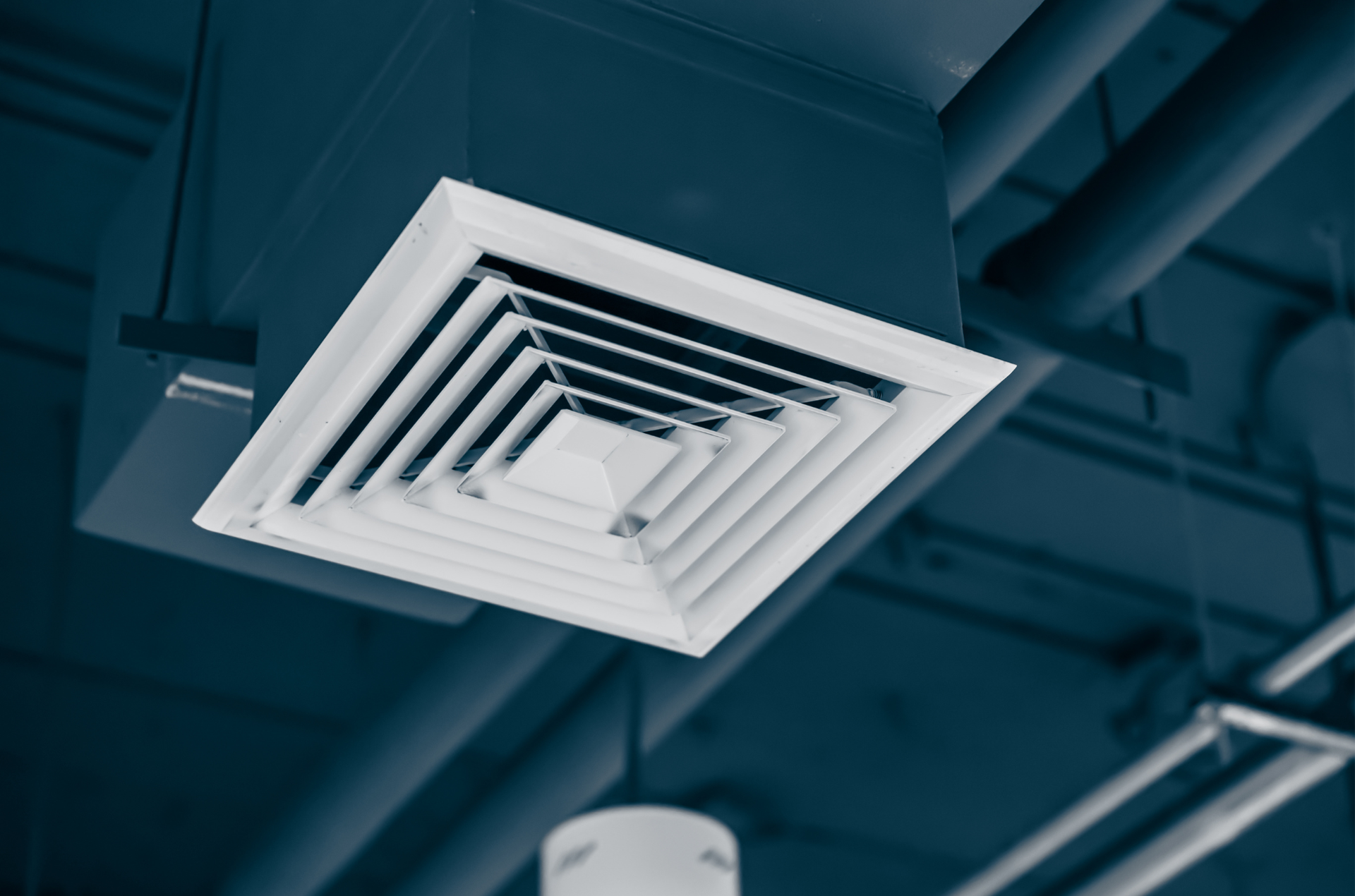 A white square ceiling air vent diffuser installed in a dark industrial-style ceiling with exposed pipes.