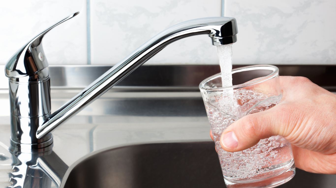 A hand holds a clear glass under a chrome kitchen faucet as water pours into the glass.