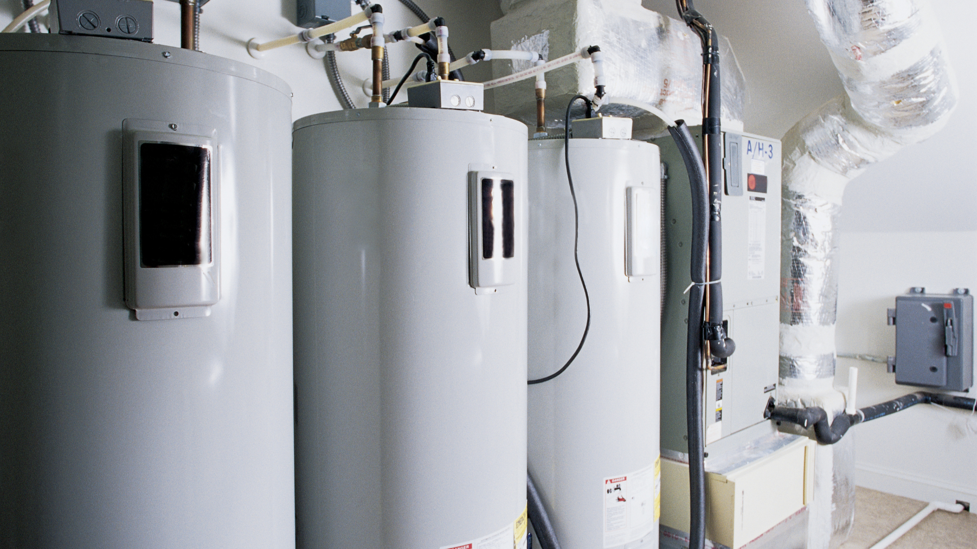 Three grey industrial water heaters arranged in a row next to mechanical utility equipment in a utility room.