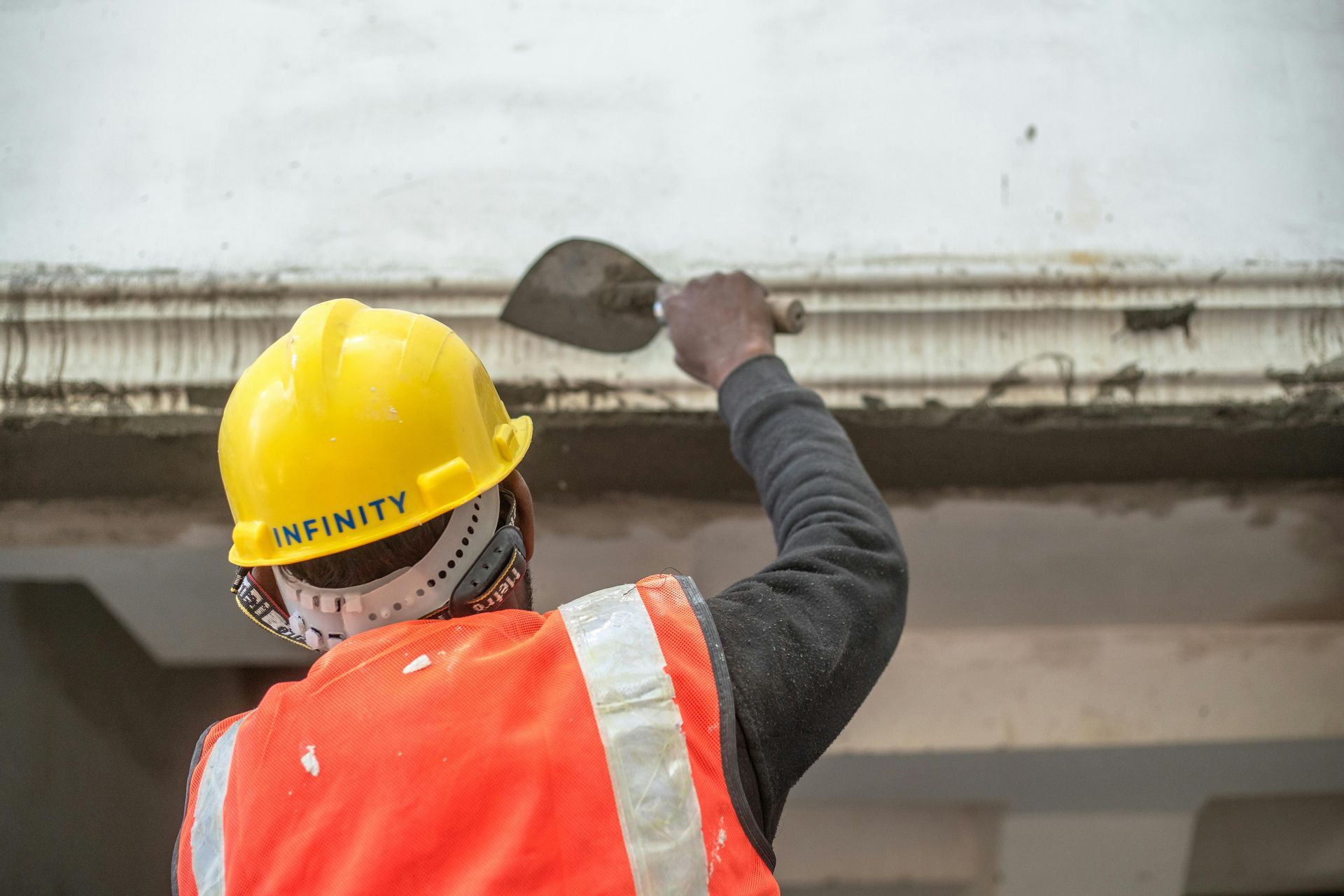A construction worker in a yellow hard hat and orange vest using a trowel to apply plaster to an overhead ledge.