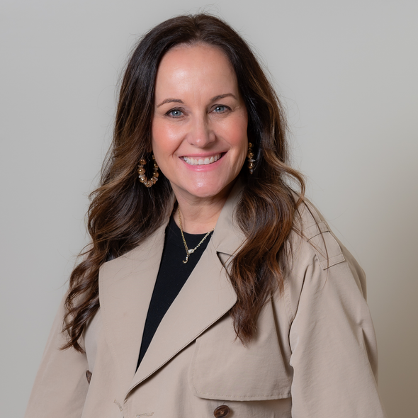 Woman with long brown hair, wearing a tan coat and black shirt, smiling at the camera.