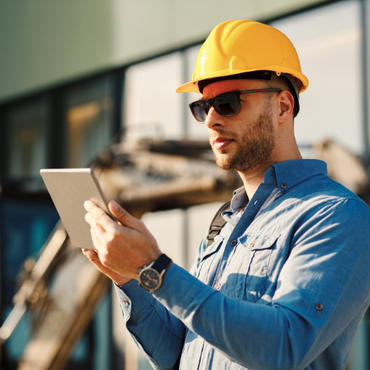 A man wearing a hard hat and sunglasses is looking at a tablet.