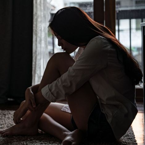 A young girl is sitting on the floor, hugging her knees, in front of a window.