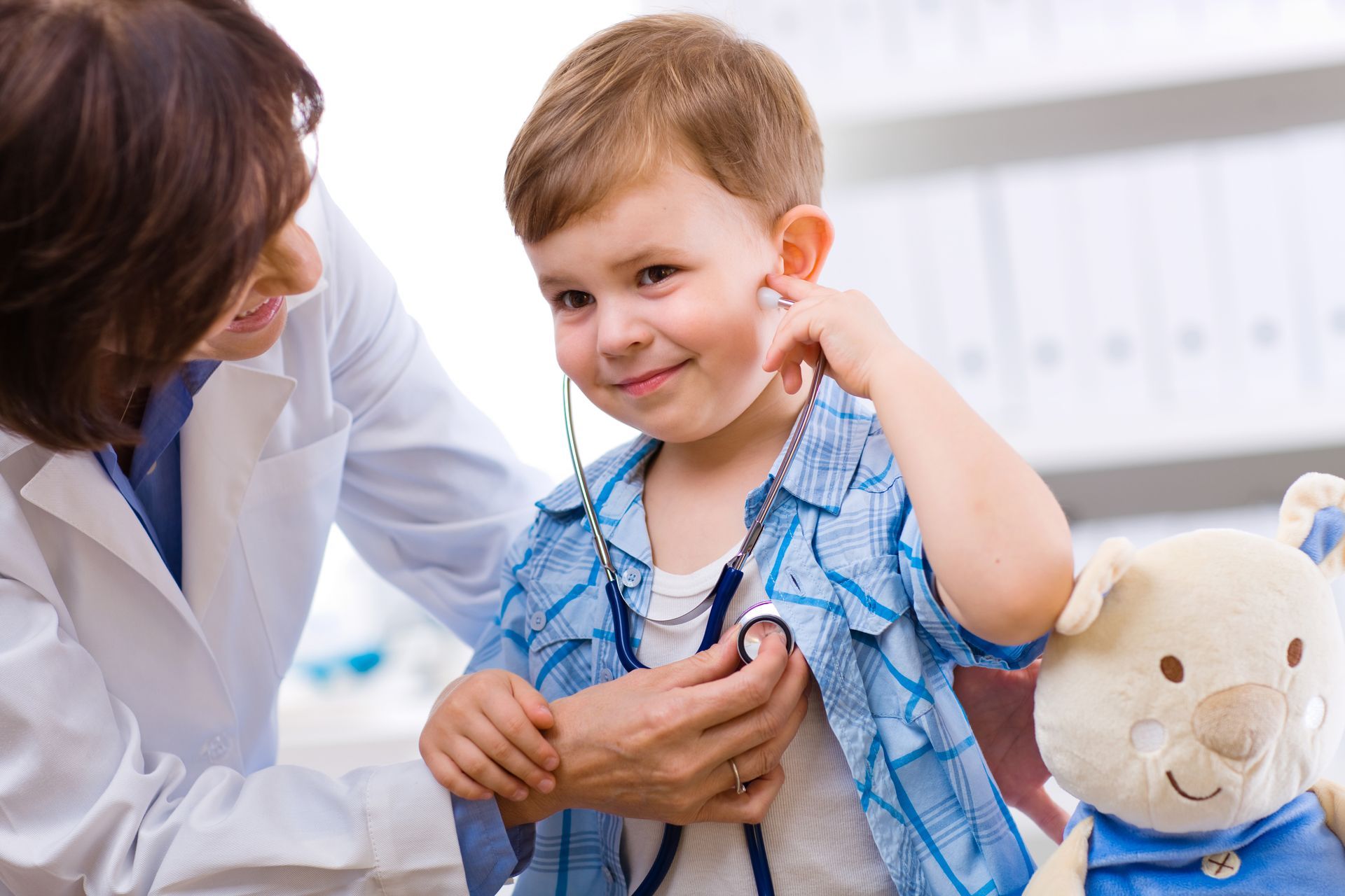 Doctor checking a smiling boy with a stethoscope, next to a stuffed animal.