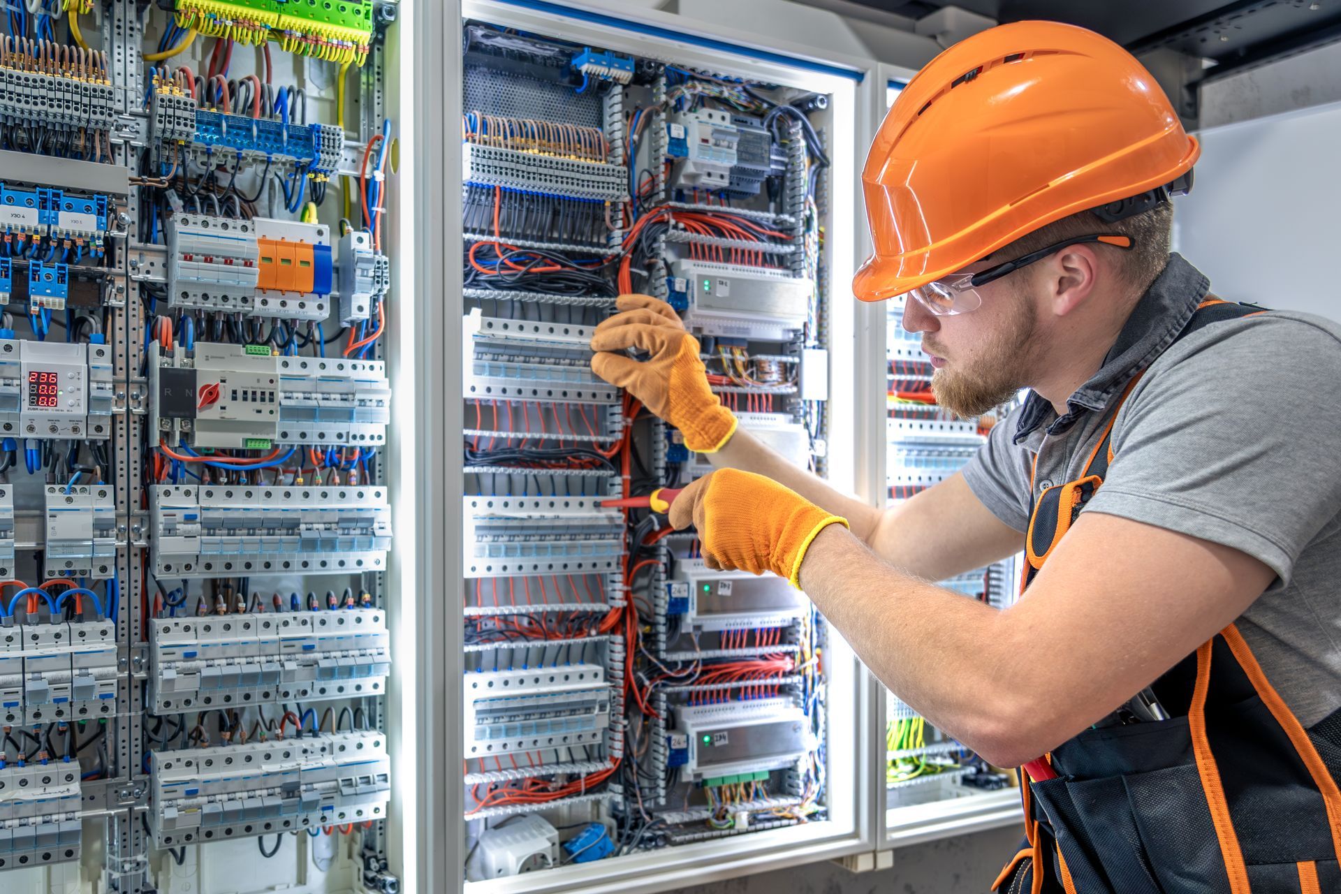 Electrician in hard hat and safety glasses, using a tablet, working on an electrical panel.