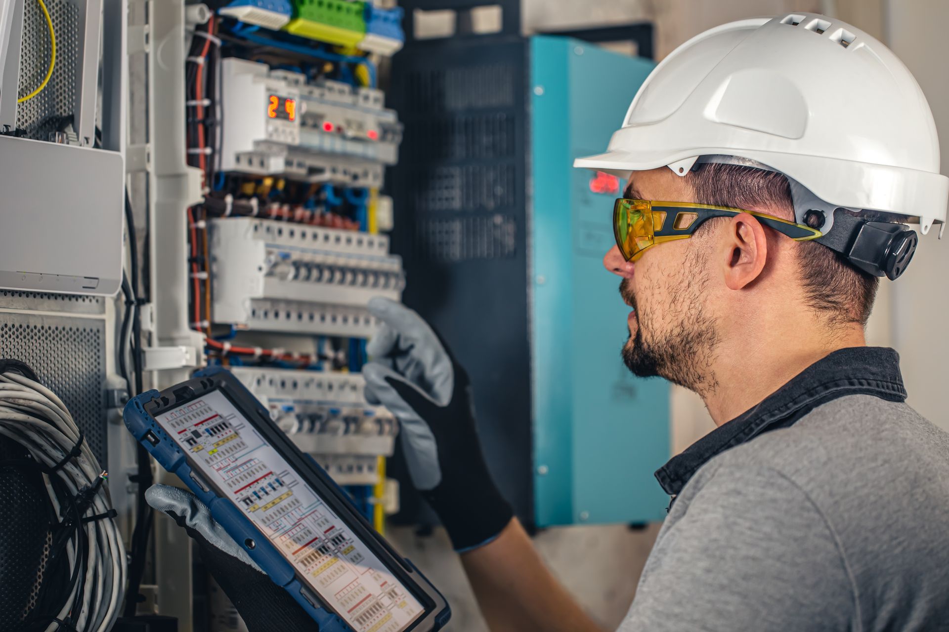 Electrician in hard hat and safety glasses, using a tablet, working on an electrical panel.
