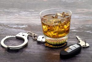 A glass of whiskey , handcuffs , and car keys on a wooden table.