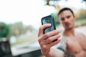 A shirtless man is taking a selfie with his cell phone.