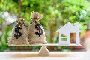 Two bags of money and a model house are on a seesaw.