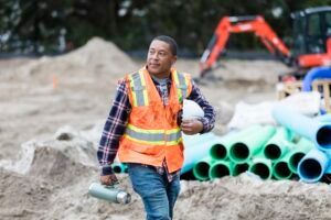 A construction worker is walking on a construction site holding a hard hat and a thermos.