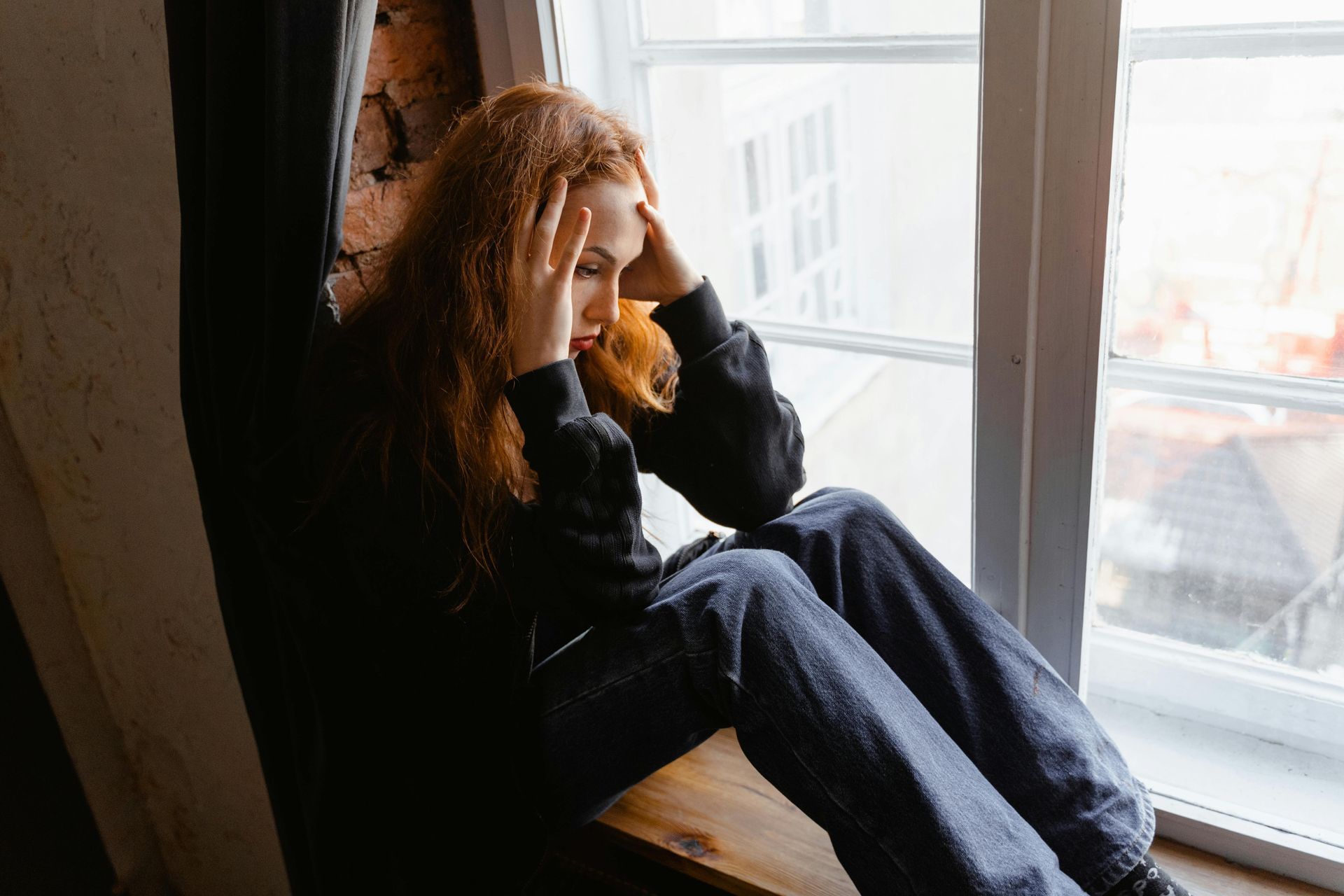 A woman is sitting on a window sill with her head in her hands.