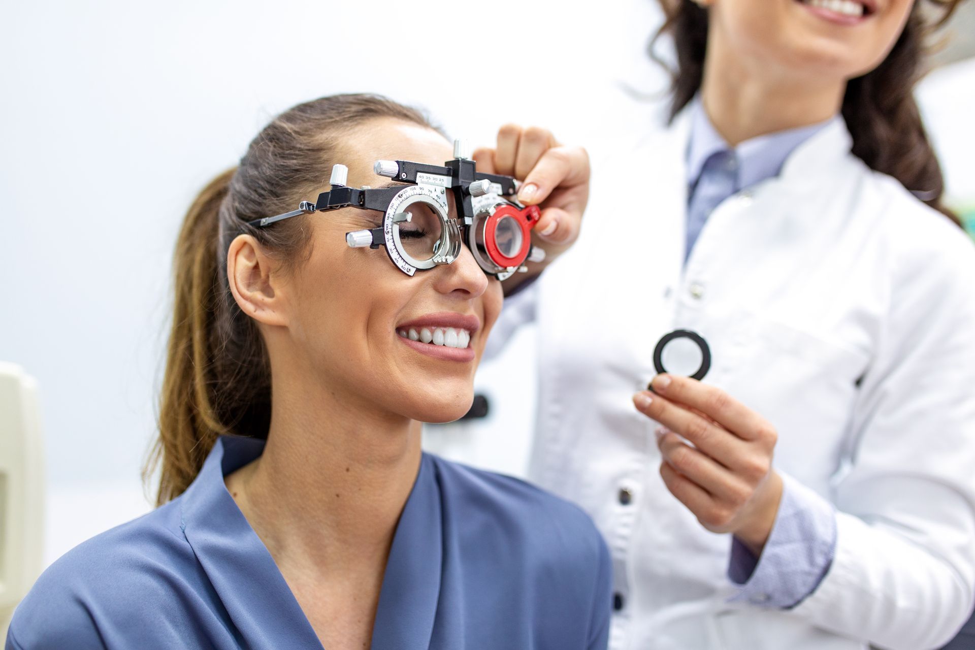 Patient wearing an eye exam phoropter while an optometrist holds a lens in a clinic