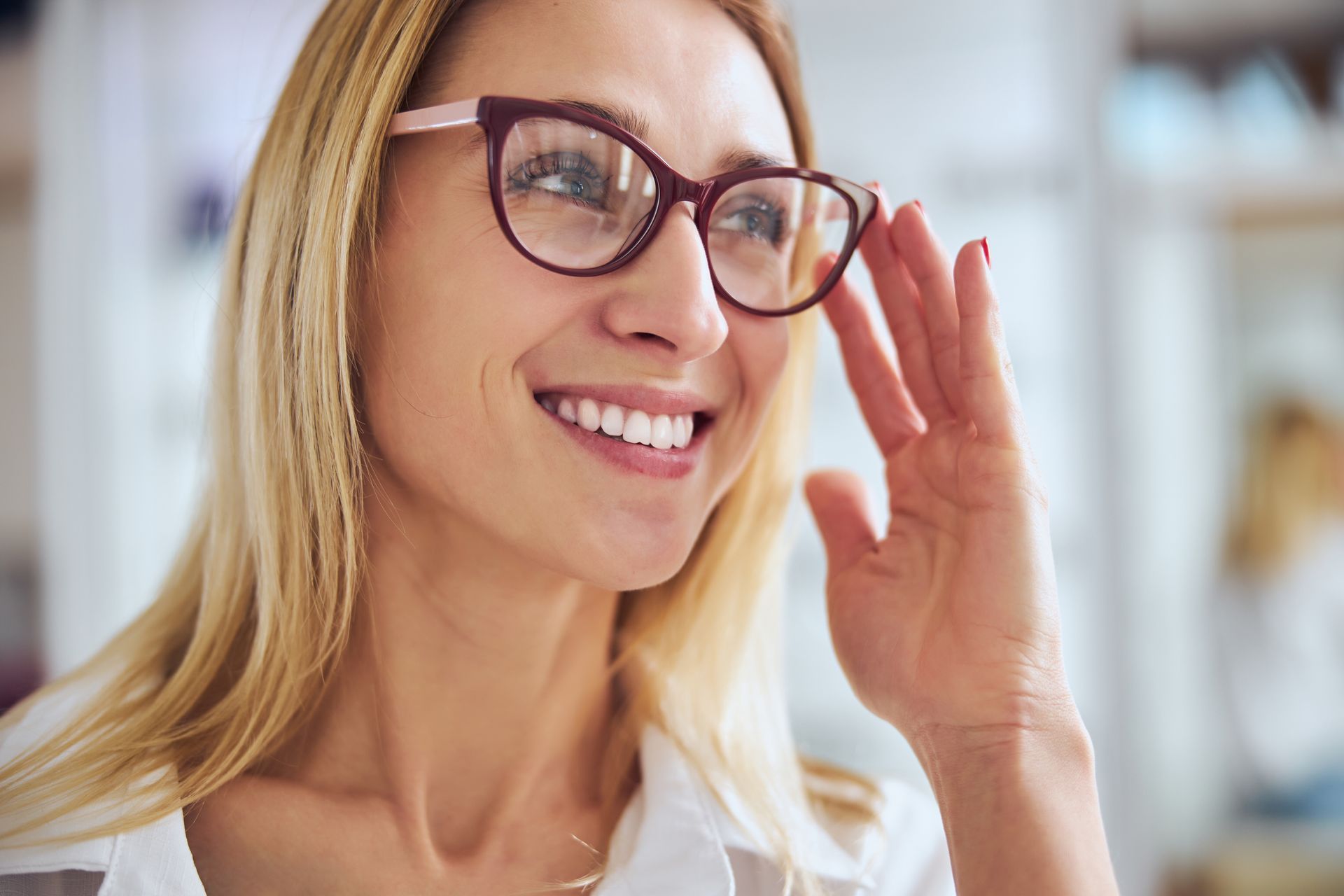 Smiling person with blonde hair adjusting brown glasses in a bright indoor setting Smiling person with blonde hair adjusting brown glasses in a bright indoor setting