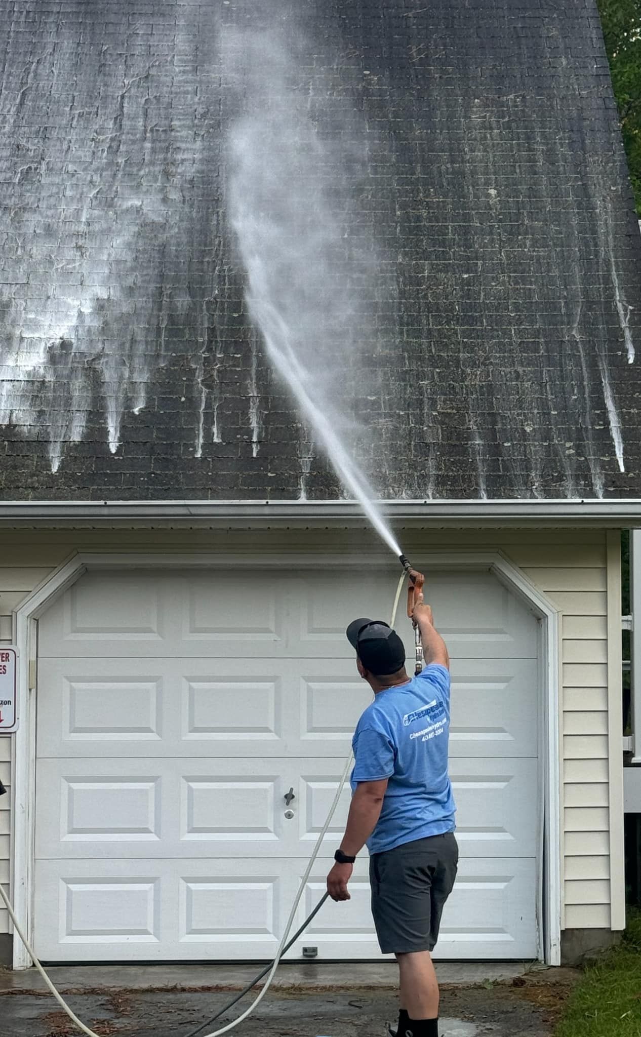 Person pressure-washes a house roof and siding with a long spray wand.