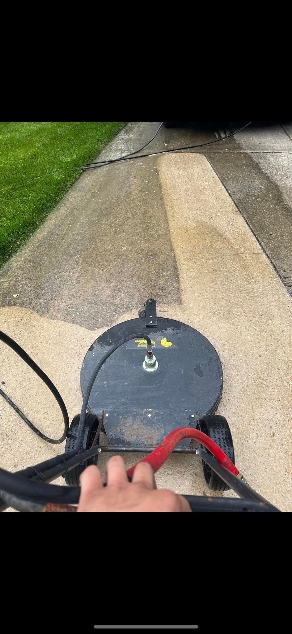 View from a lawn mower on a driveway beside grass, with a hand on the handle.