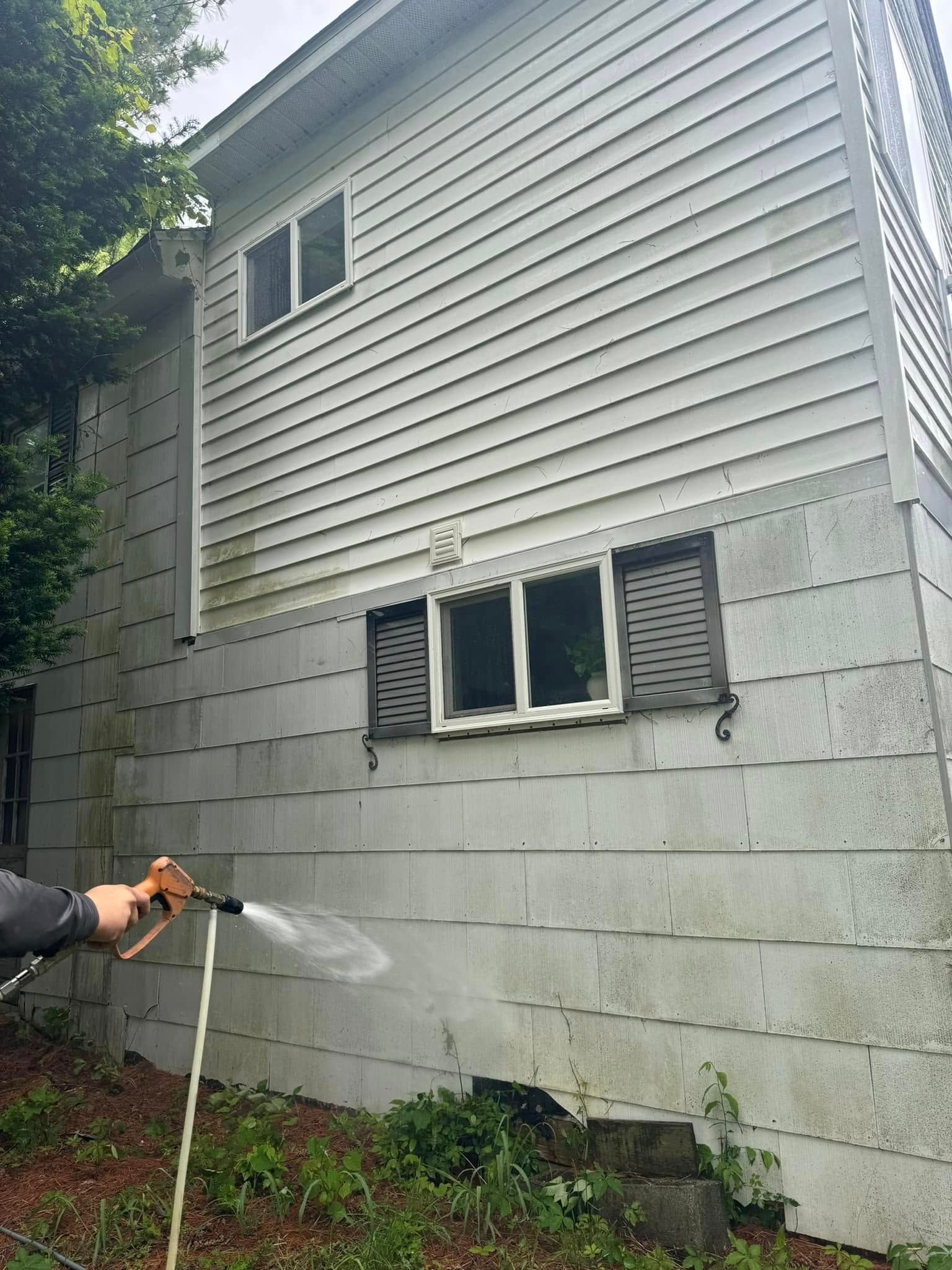 Person spraying water on a house’s lower cinder block wall and white siding with a hose.