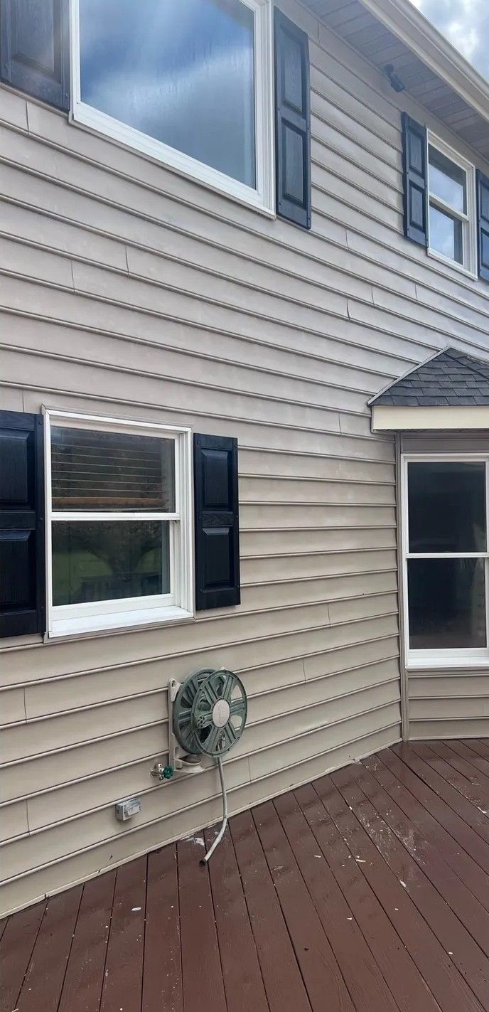 Exterior of a beige house with white shutters, two windows, a wall-mounted hose reel, and a brown deck railing