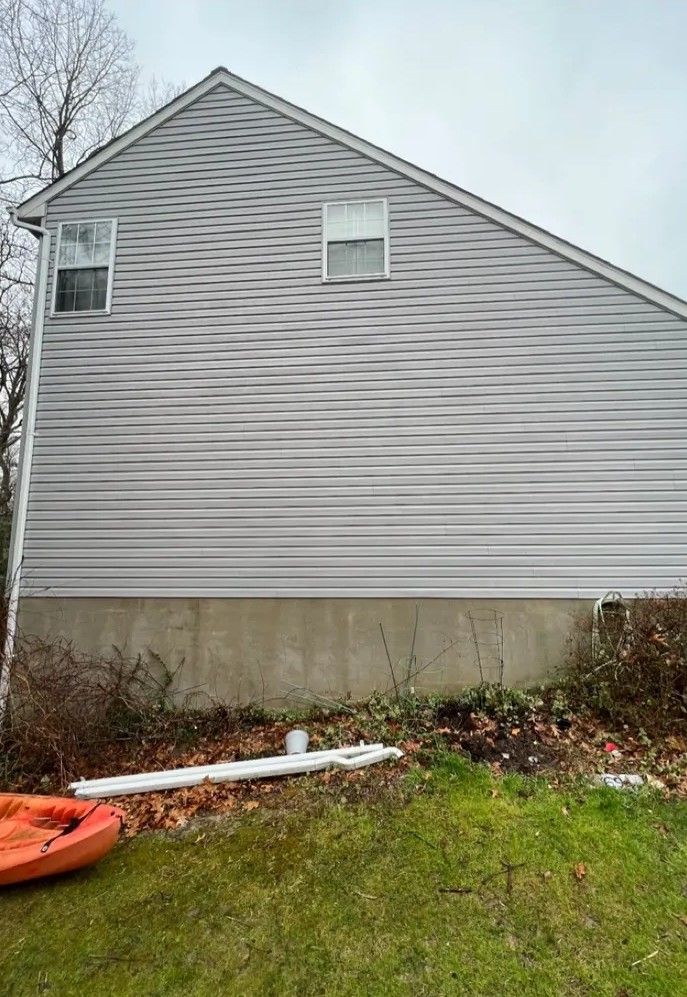 Side view of a gray house with horizontal siding, small windows, and a concrete foundation overgrown with shrubs.