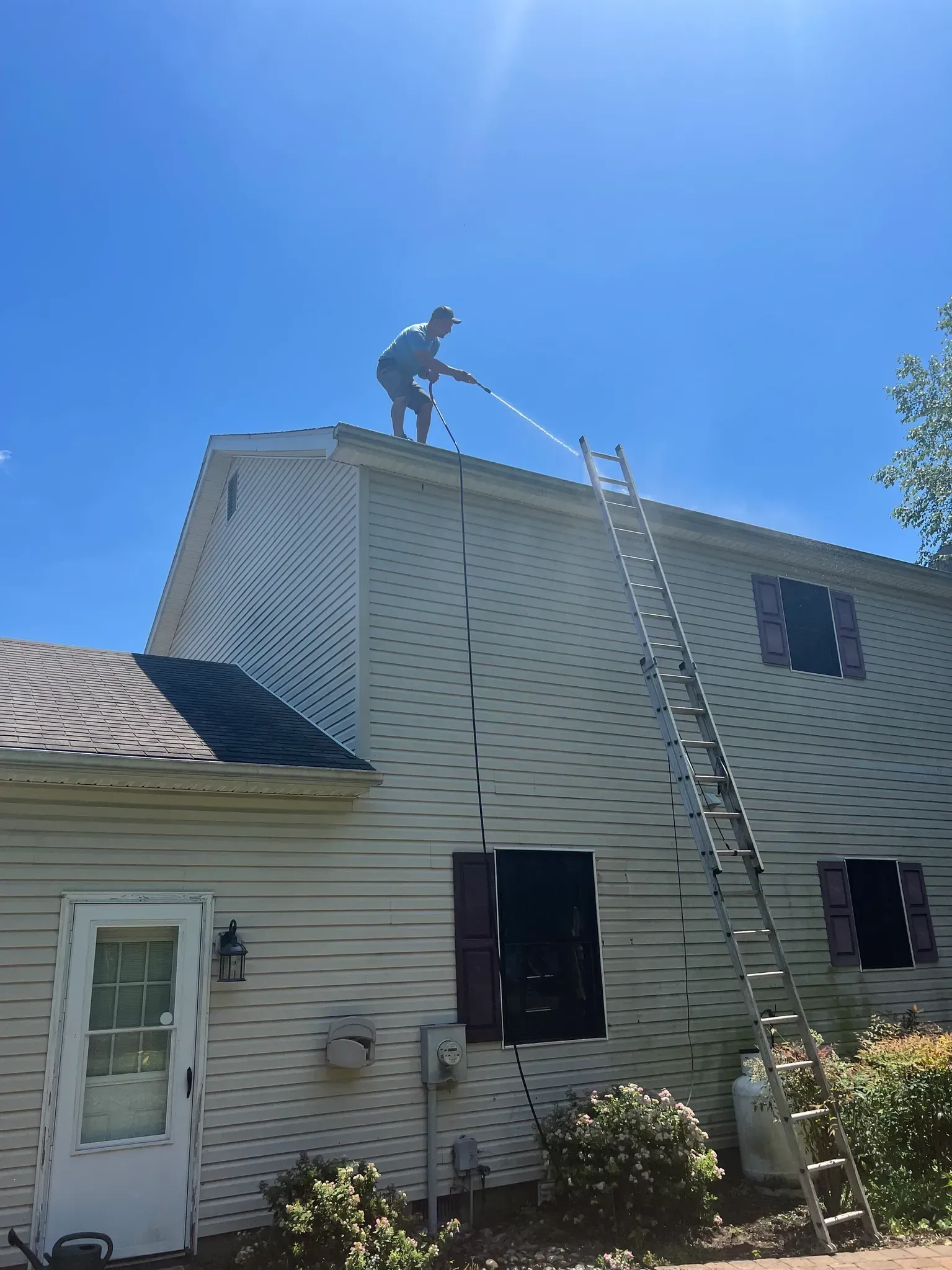 Person on a roof spraying water near a house, with a ladder leaning against the siding.