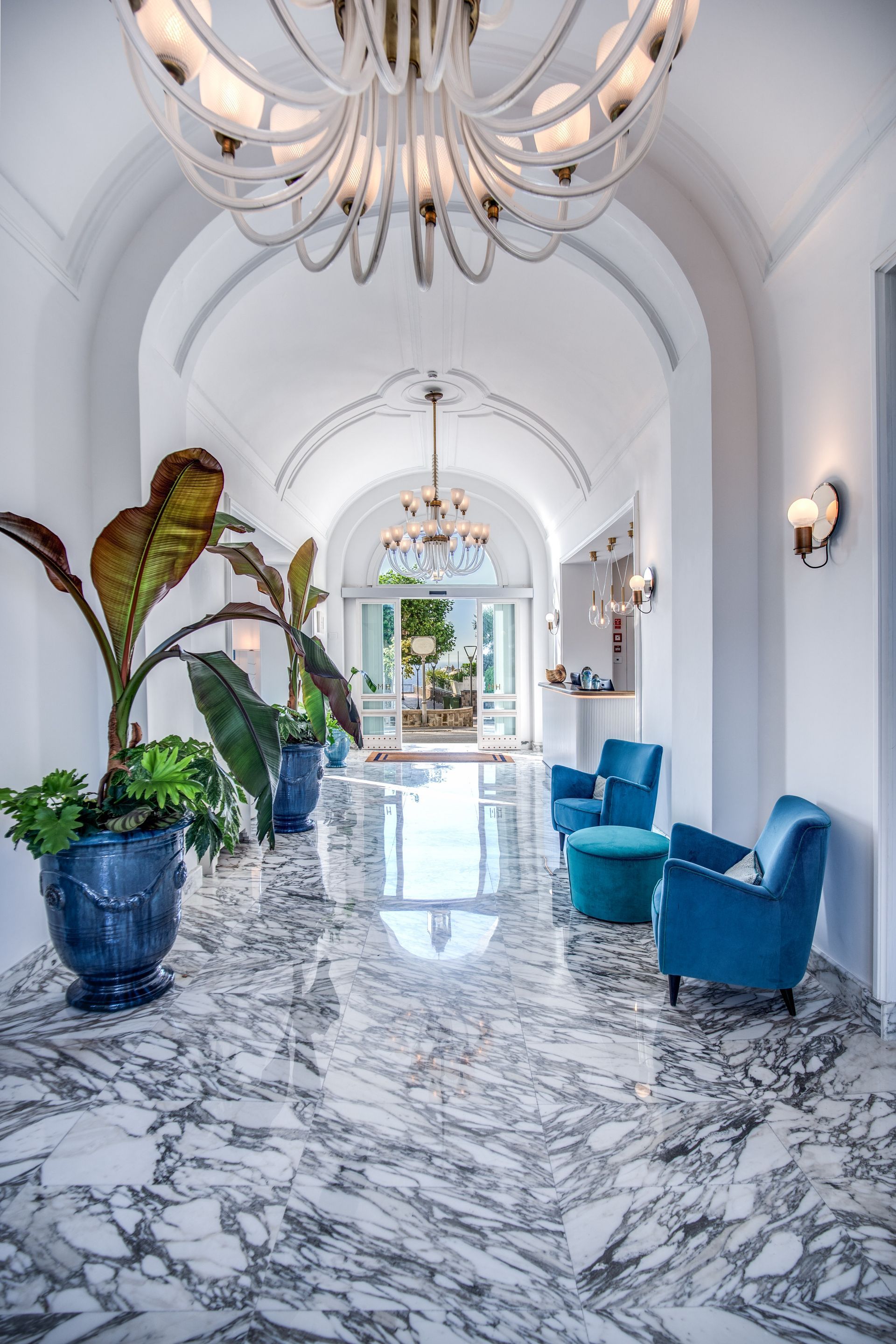 A marble-floored hotel hallway with white arched ceilings, blue armchairs, and large potted plants under crystal chandeliers.
