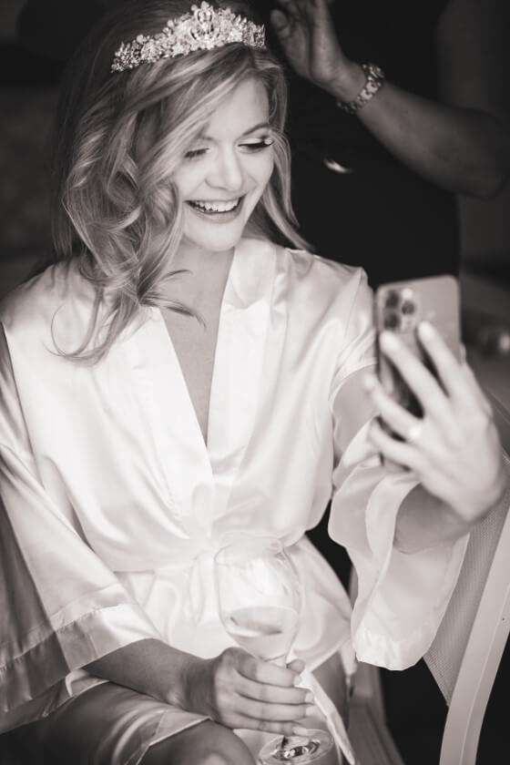 A bride wearing a tiara and silk robe smiles while taking a selfie with a smartphone and holding a glass of wine.