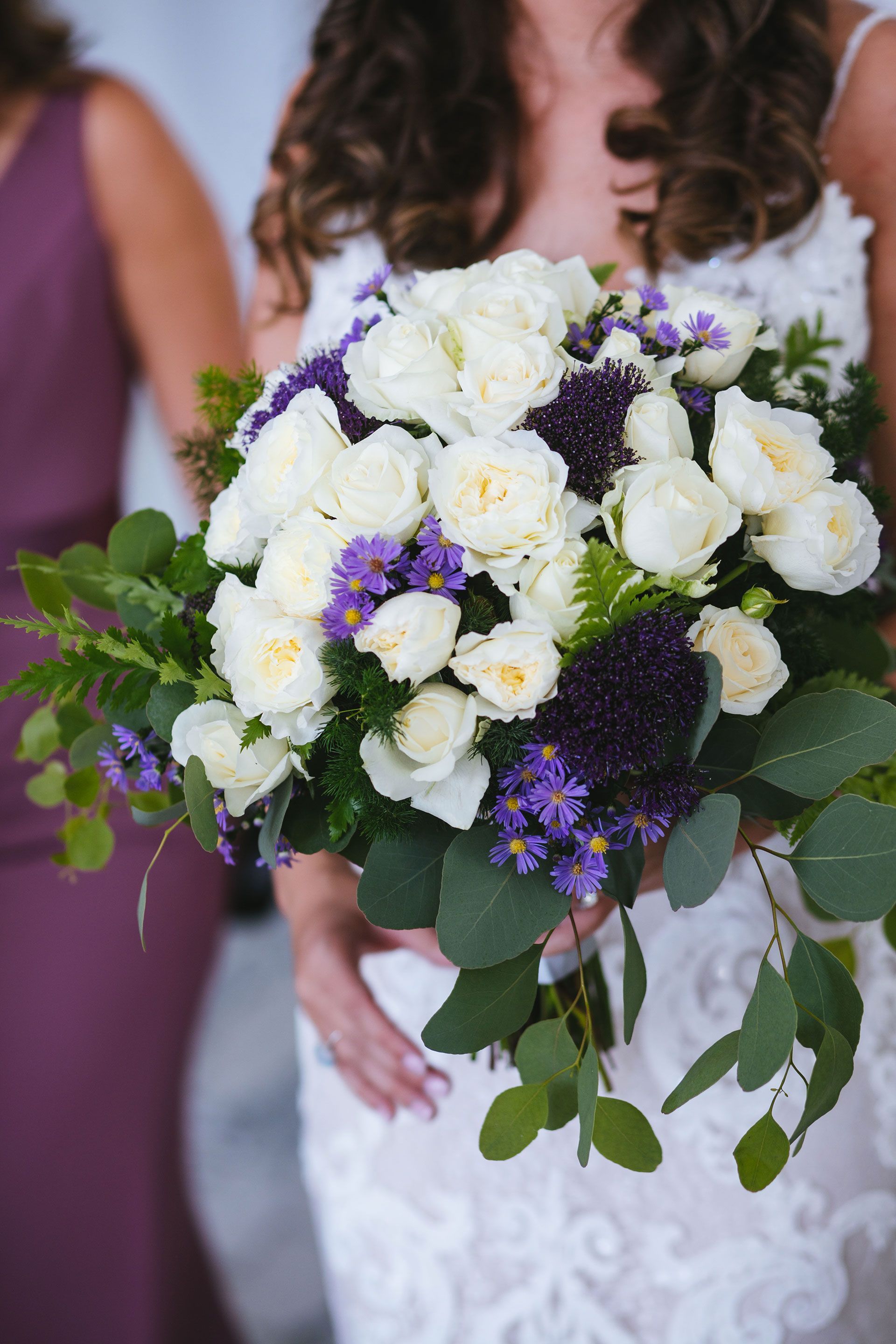 A bride in a white lace dress holds a bridal bouquet of white roses, deep purple flowers, and eucalyptus greenery.