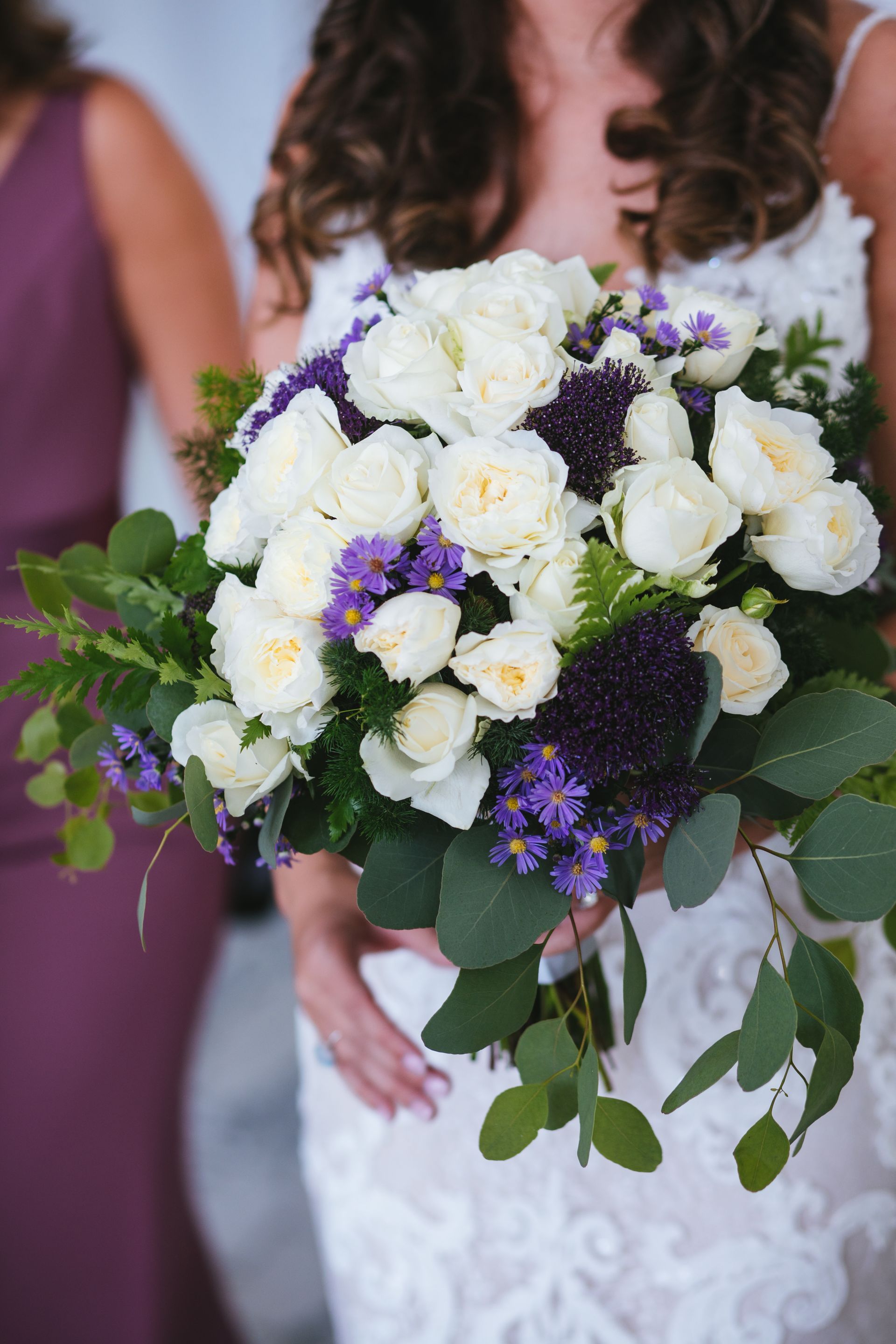 A bride holds a wedding bouquet of white roses, deep purple flowers, and eucalyptus greenery, with a bridesmaid nearby.