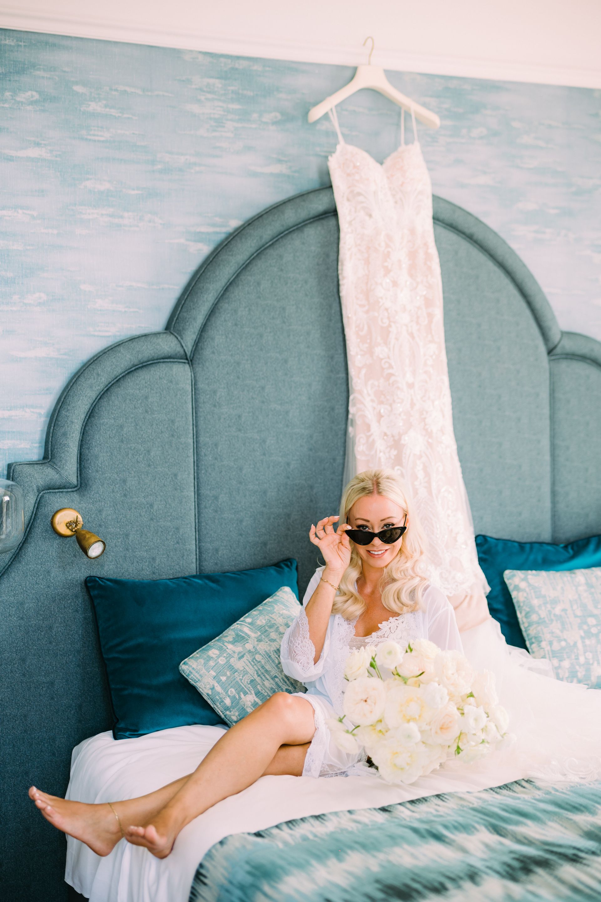 A person in a white robe and sunglasses sits on a teal bed, holding flowers, with a wedding dress hanging above.