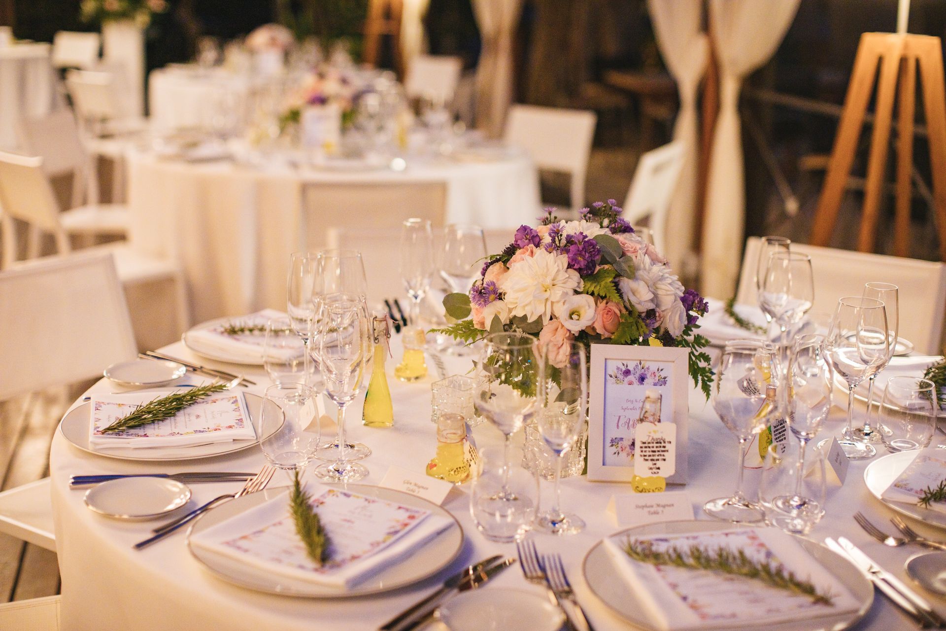 Tables set for a wedding reception with white linens, floral centerpieces, glassware, and sprigs of greenery on napkins.