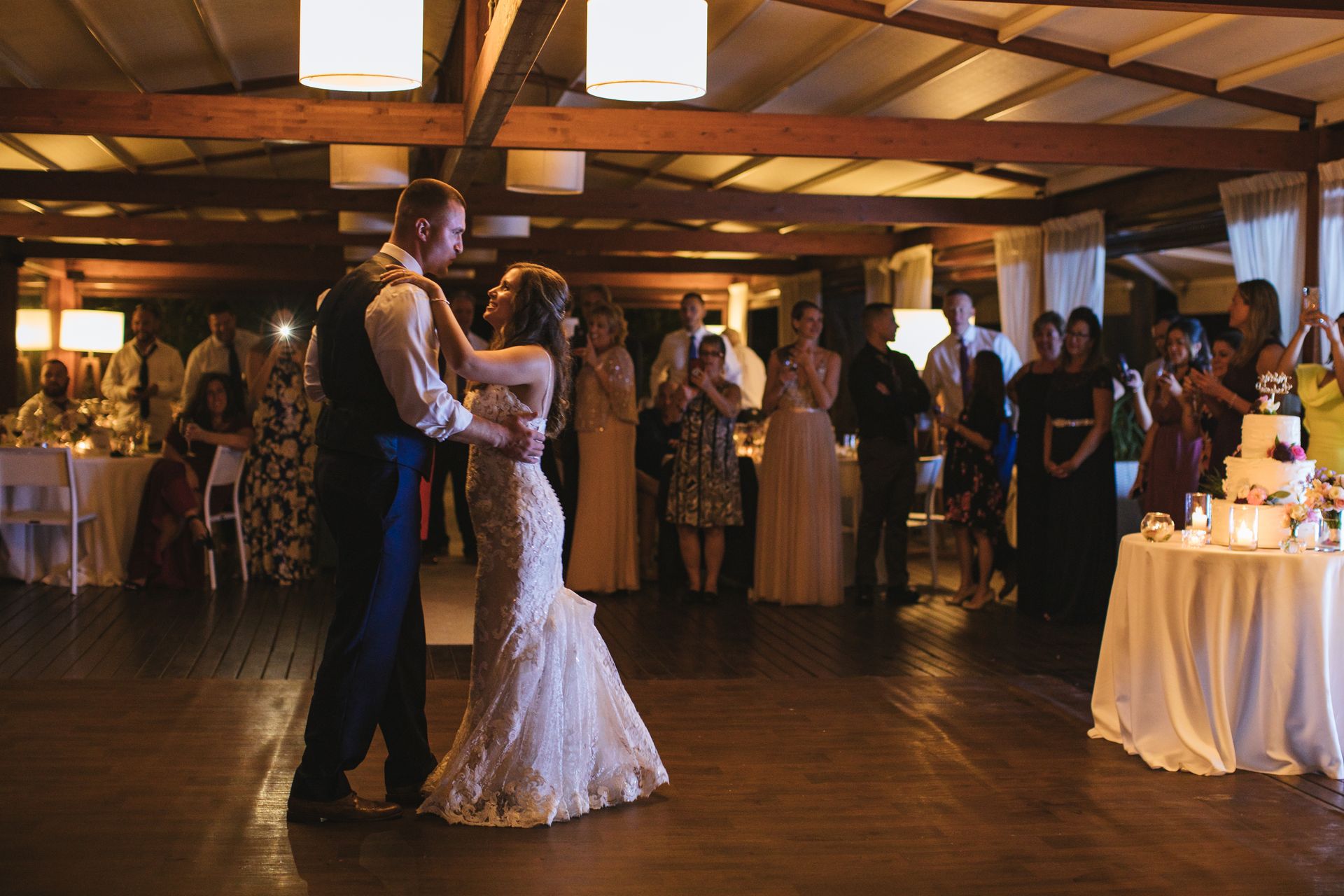 A couple dancing at their wedding reception in a dimly lit, rustic venue with guests watching in the background.