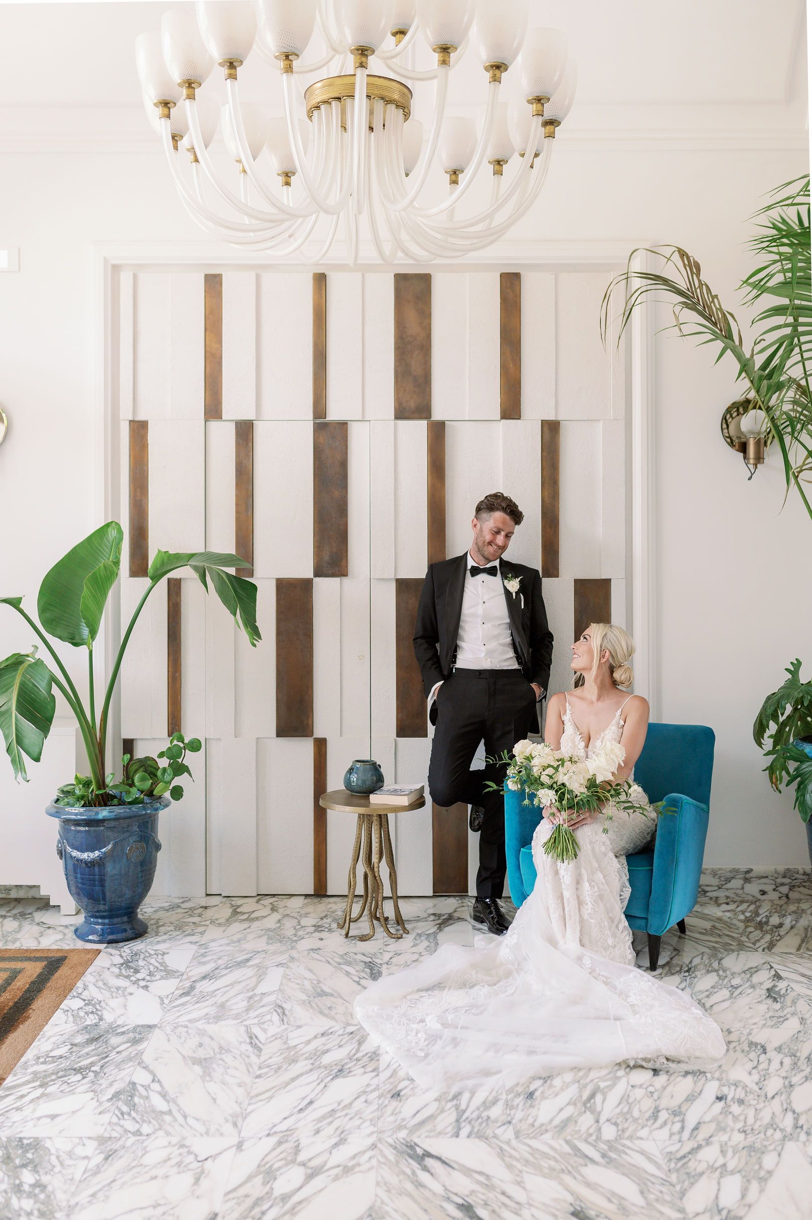 Una sposa in abito bianco siede su una sedia di velluto blu accanto allo sposo in smoking nero, davanti a una parete moderna e geometrica.
