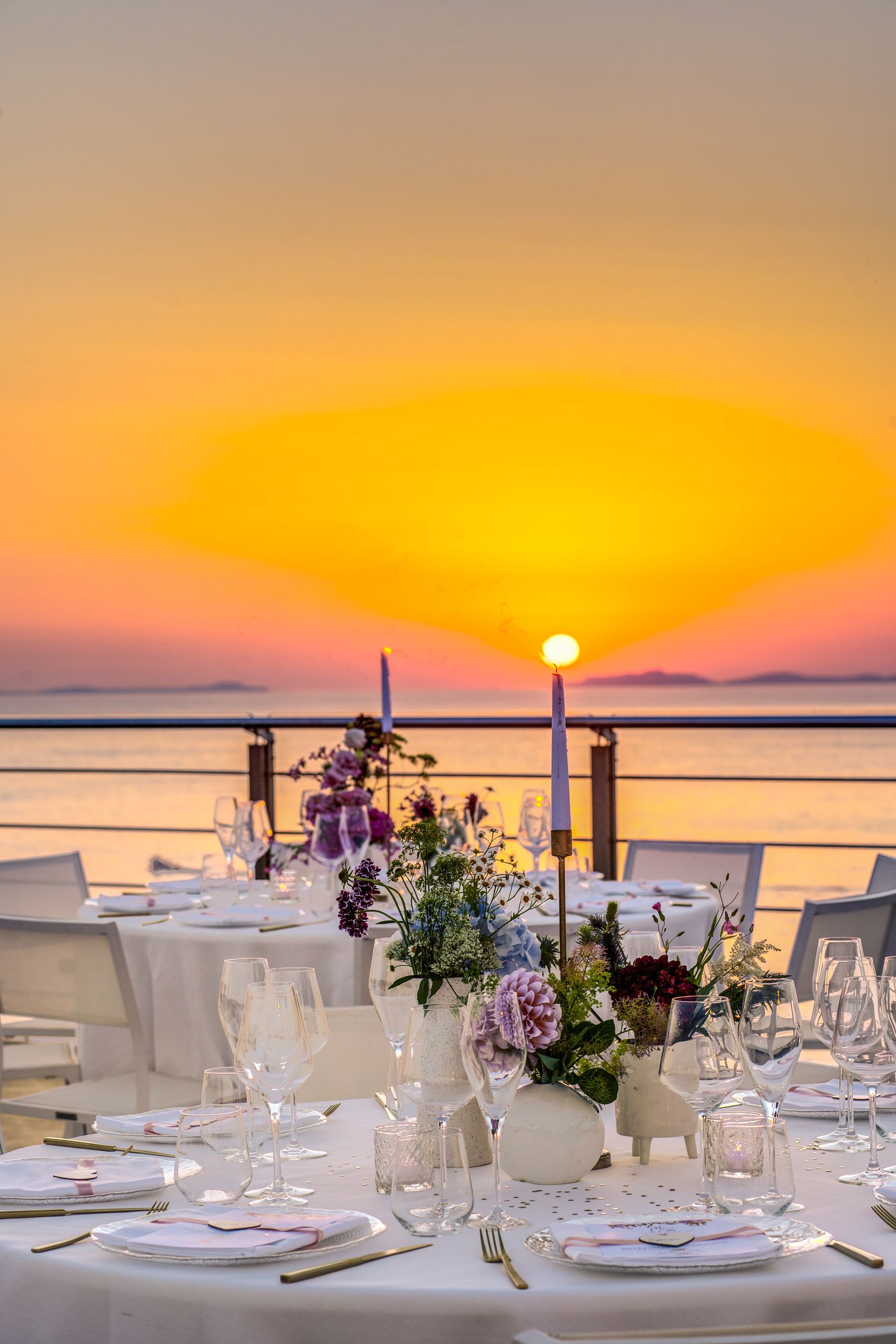 Tables set for dinner on an outdoor balcony overlooking a scenic ocean sunset with vibrant orange and yellow hues.