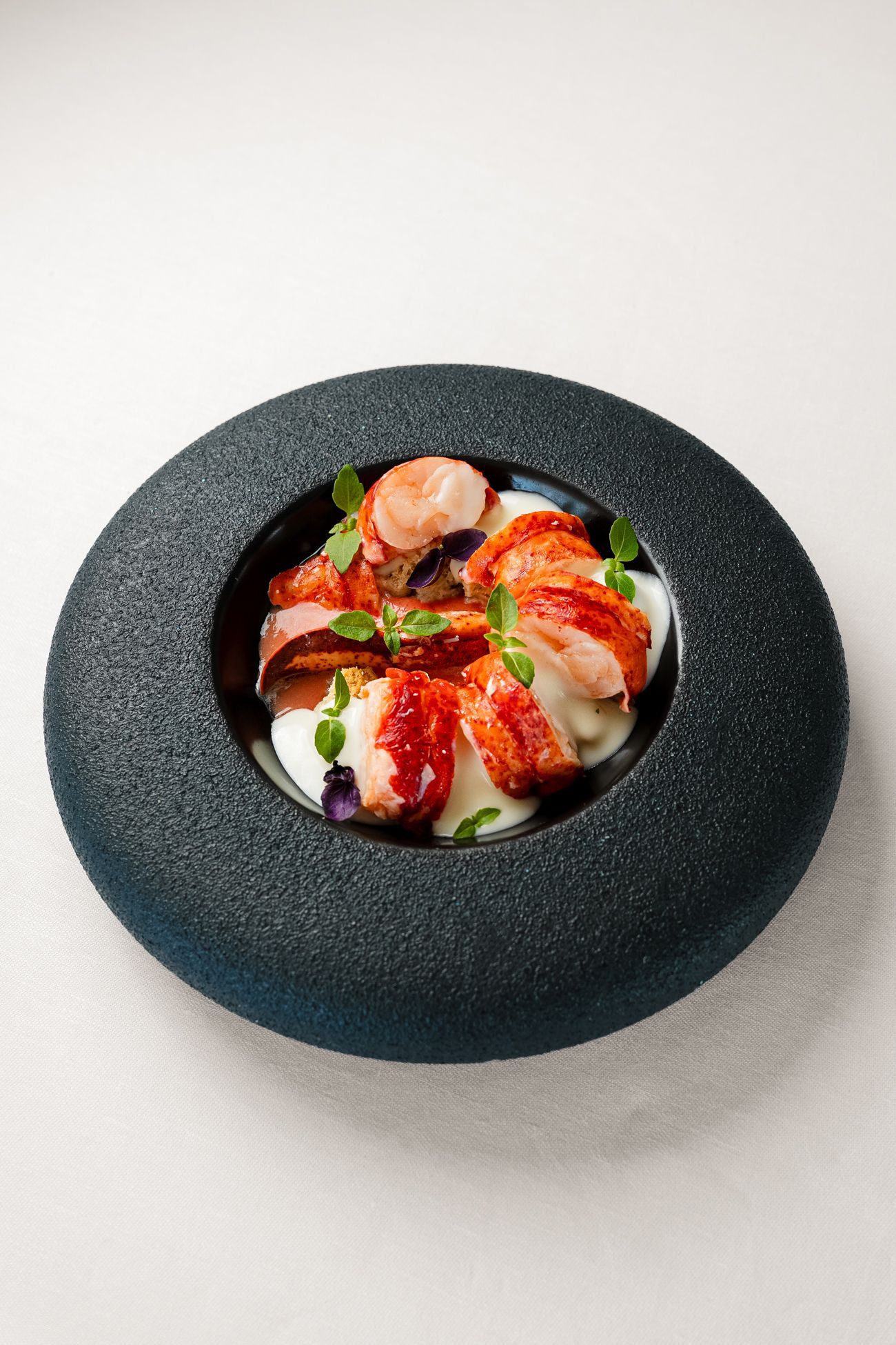 Pezzi di astice cotto serviti in una salsa bianca cremosa, guarniti con erbe aromatiche in una ciotola scura e dalla superficie ruvida, simile alla pietra.