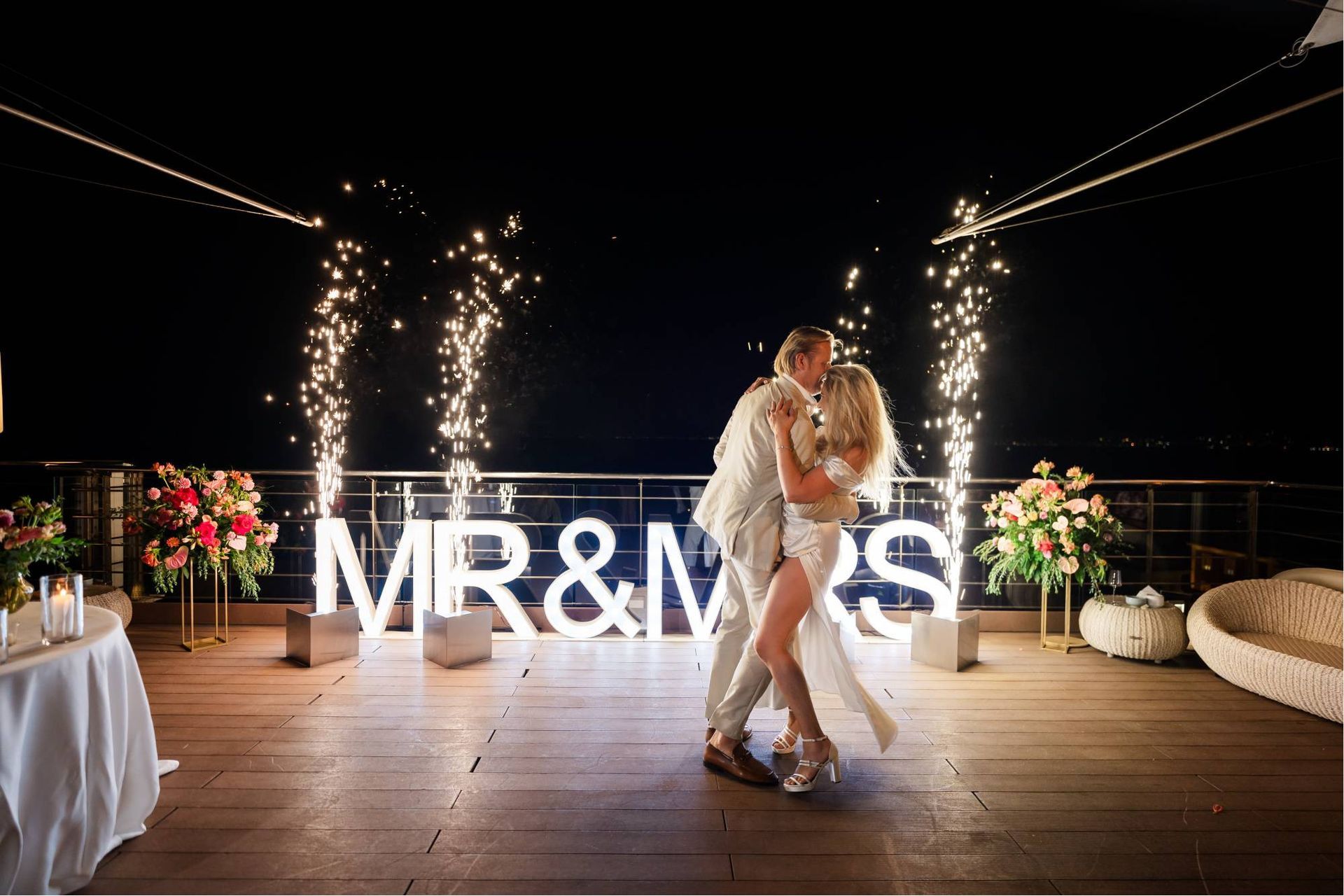 A couple embraces and dances on an outdoor deck at night, framed by sparklers and a glowing 