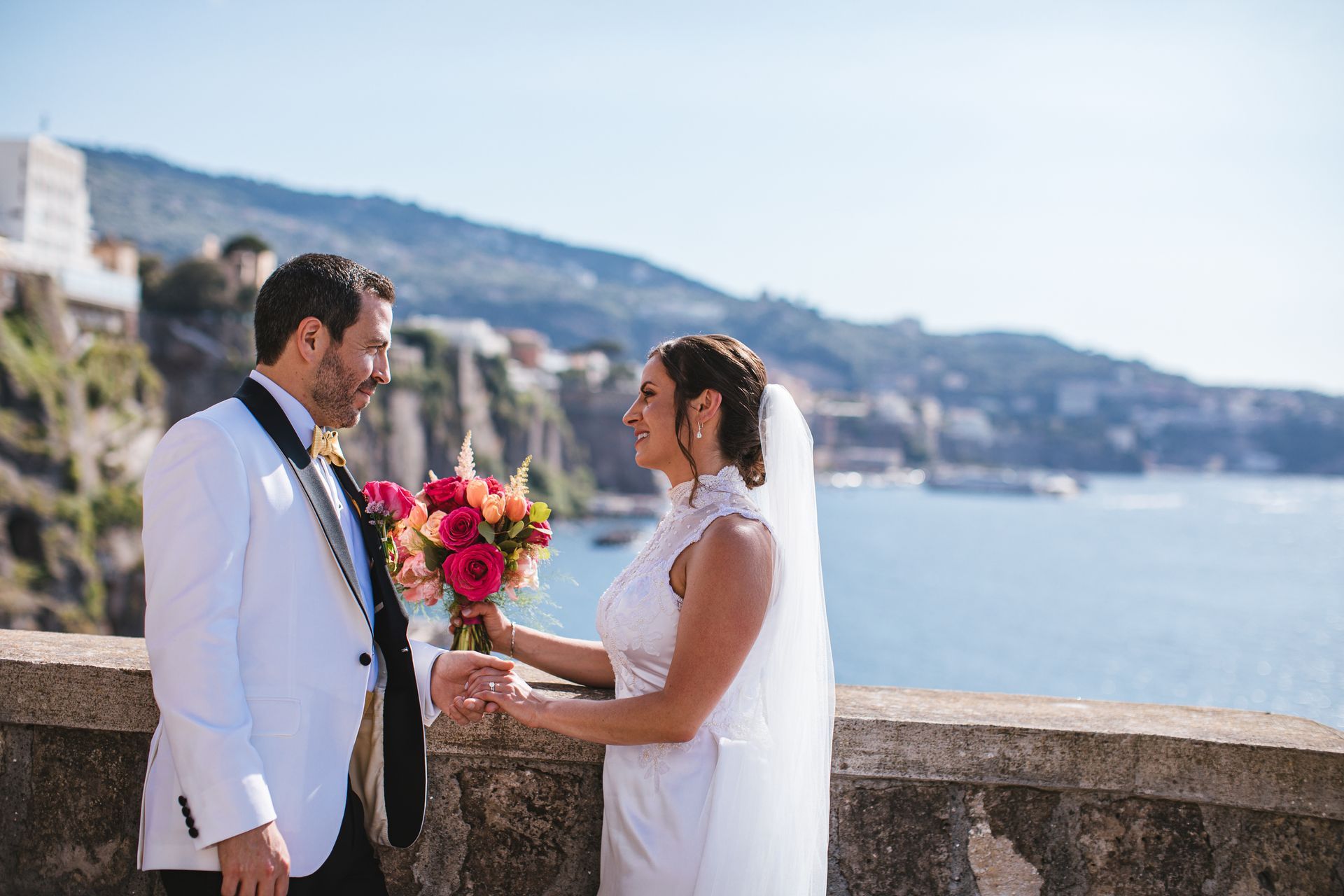 A bride and groom in wedding attire hold hands and look at each other on a balcony overlooking the sea and a cliffside.