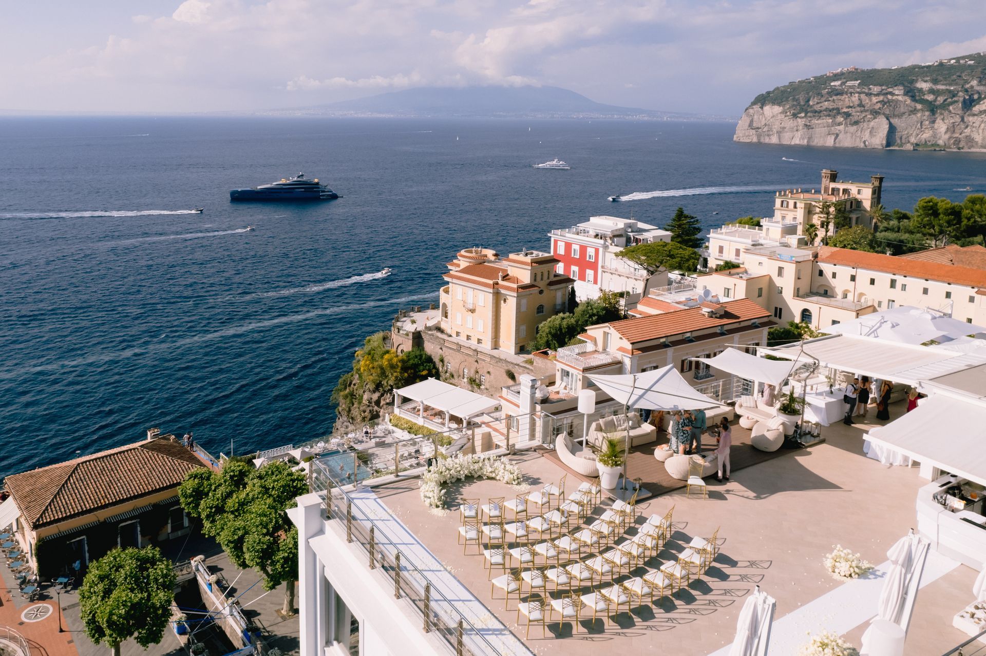 A high-angle view of a seaside wedding ceremony setup with white chairs on a terrace overlooking the blue ocean.