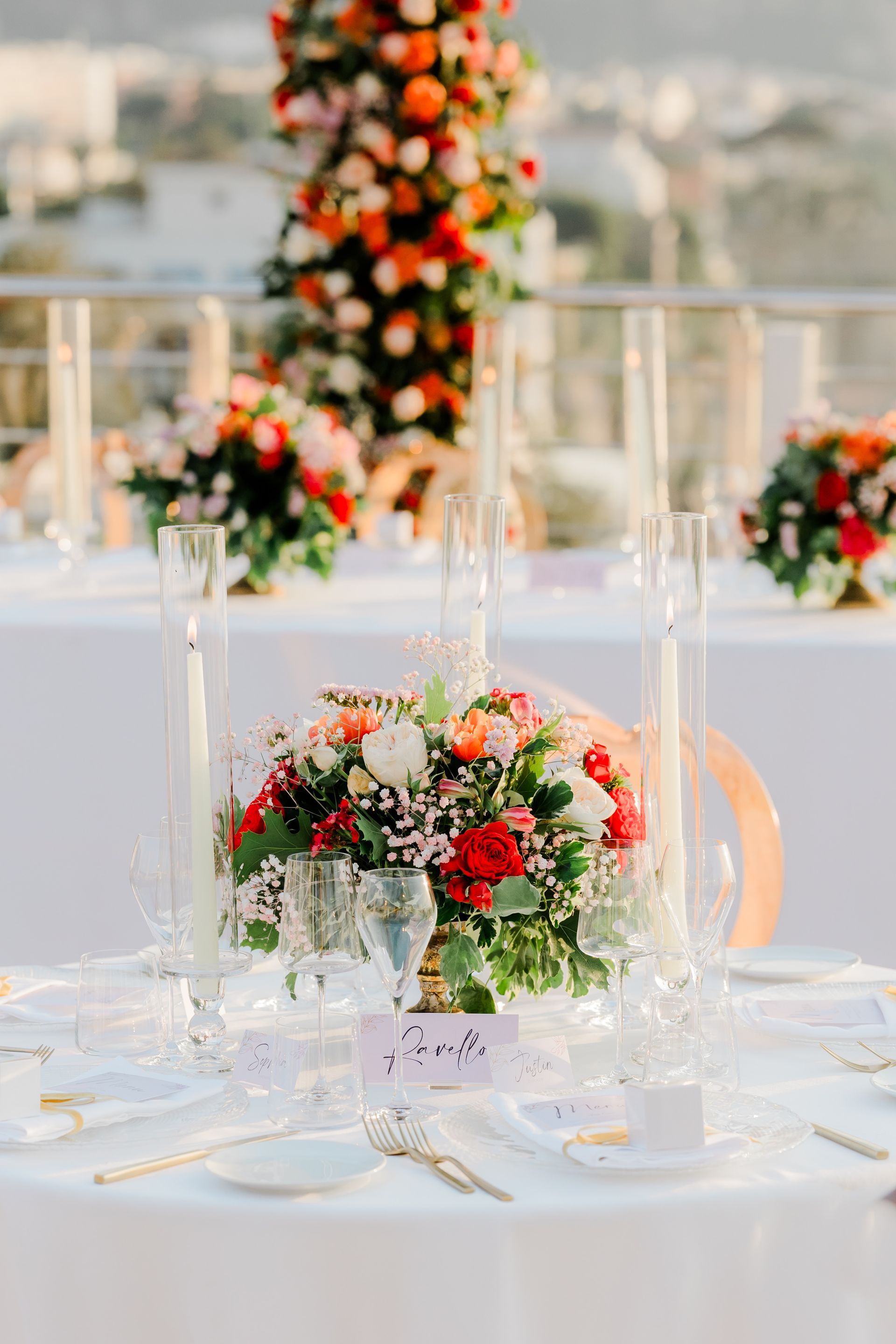 A round outdoor table set for a wedding with a central floral centerpiece, tall glass candle holders, and white linens.