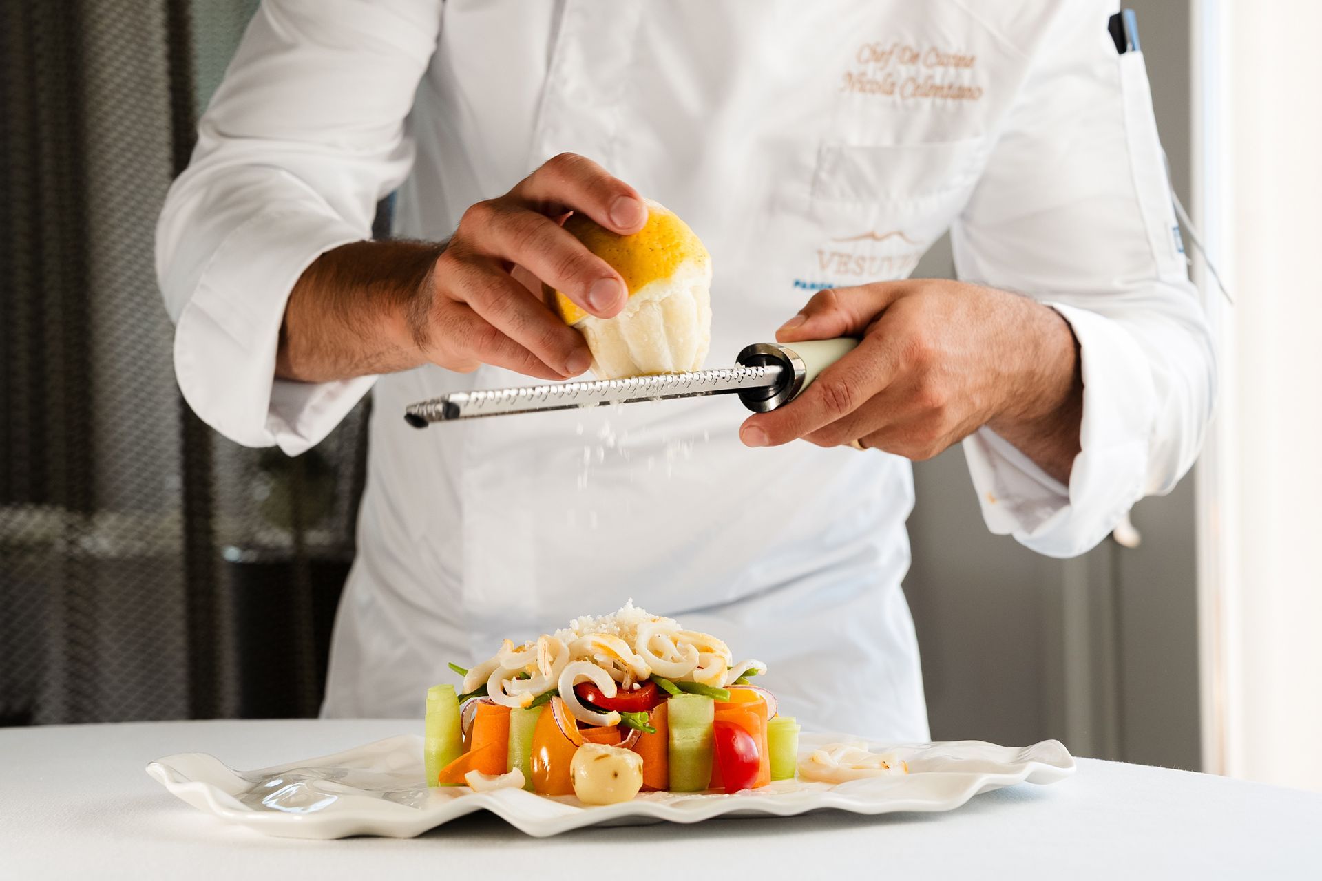 Uno chef in uniforme bianca usa una grattugia per agrumi su un colorato piatto di verdure adagiato su un piatto bianco.