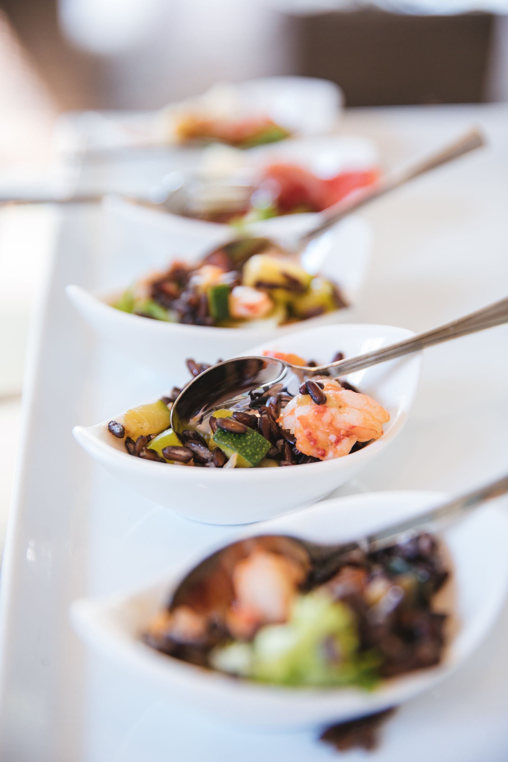 A row of white ceramic spoons on a long platter, each filled with a small appetizer of dark rice, salmon, and vegetables.