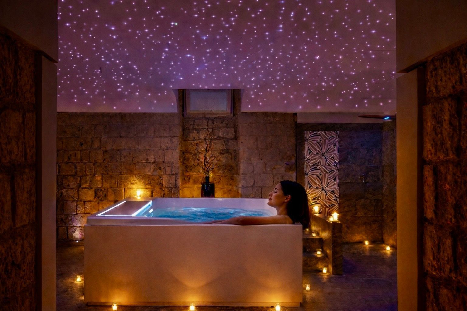 Una persona si rilassa in una vasca idromassaggio illuminata, all'interno di una stanza in pietra, sotto un soffitto scuro illuminato da piccole luci scintillanti.