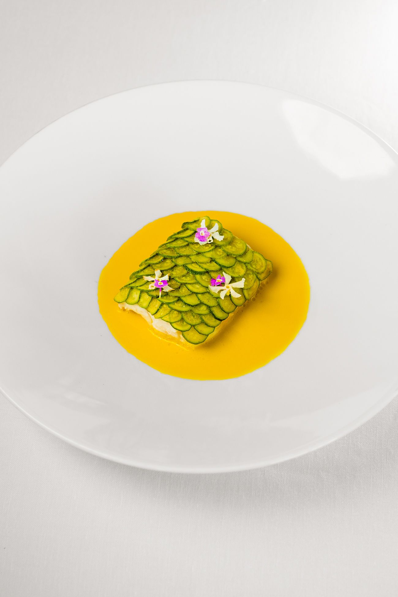 Un pezzo di pesce rettangolare sormontato da fette di cetriolo sovrapposte e piccoli fiori edibili, adagiato su una salsa gialla.
