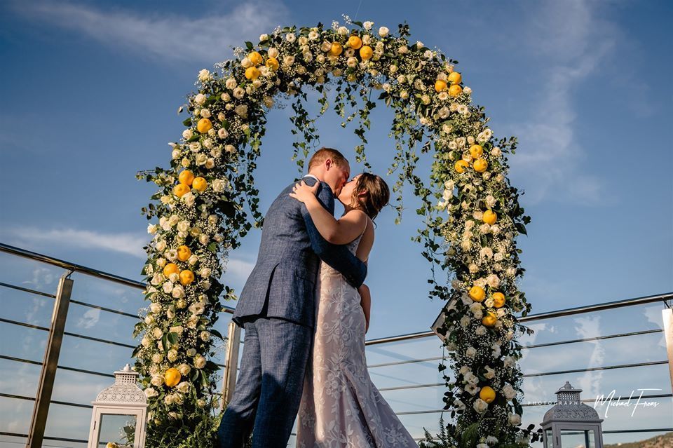 A bride and groom in wedding attire kiss under a floral arch decorated with white flowers and lemons against a blue sky.