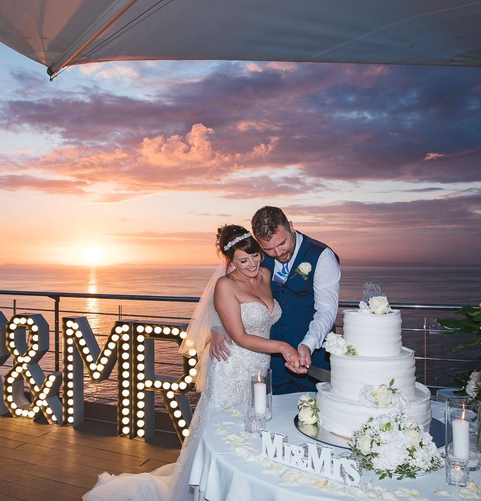 A bride and groom cut a multi-tiered white wedding cake on an outdoor balcony at sunset overlooking the ocean.