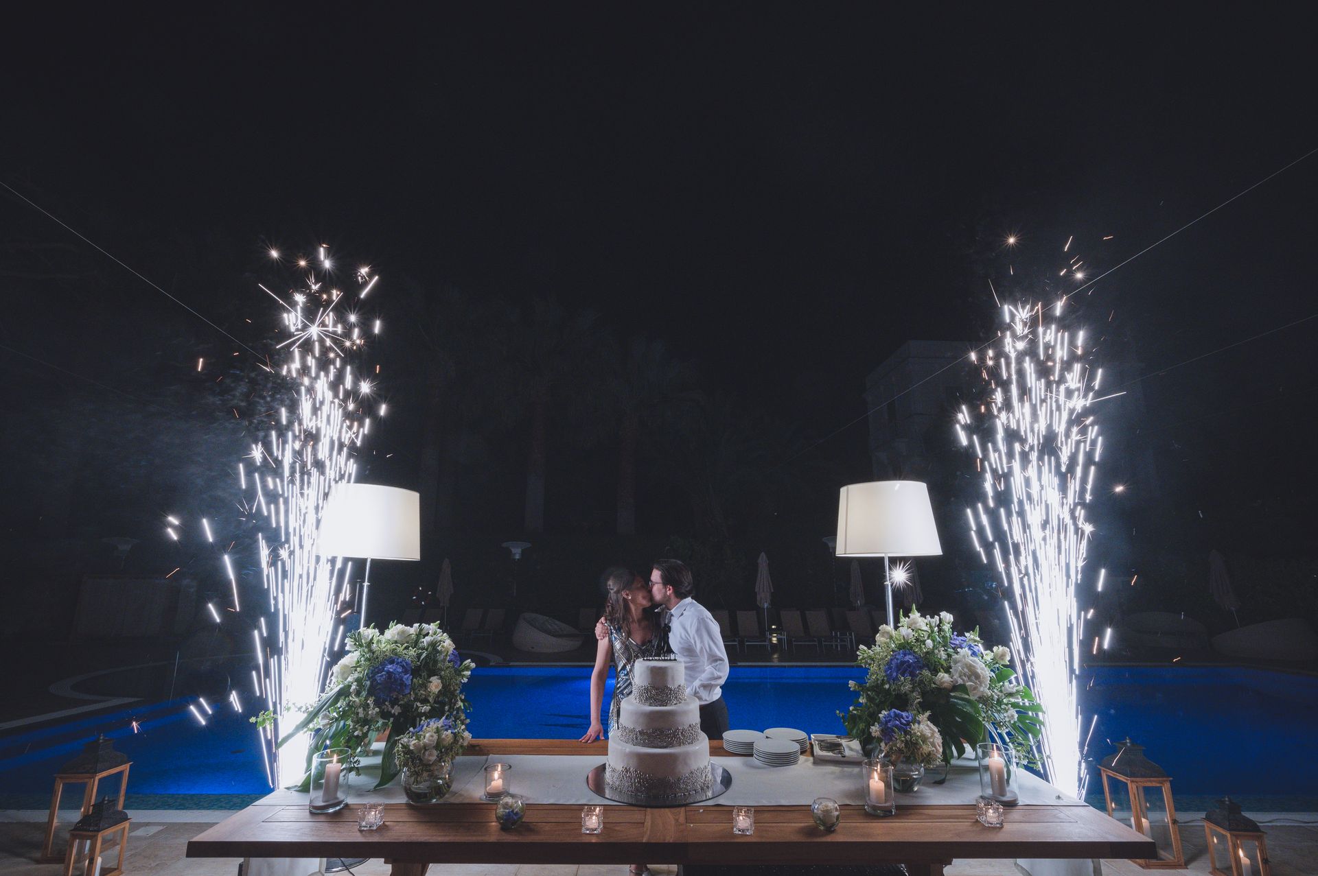 A couple stands behind a tiered wedding cake at night by a pool, framed by two vertical fireworks fountains and lamps.