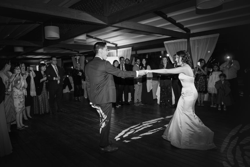 A bride and groom share a dance in a dimly lit, crowded venue, holding hands as they move across a dark wooden floor.