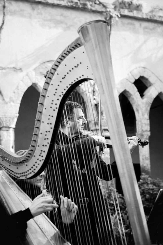 A musician plays a violin behind the strings of a large harp, set against a backdrop of arched stone architecture.