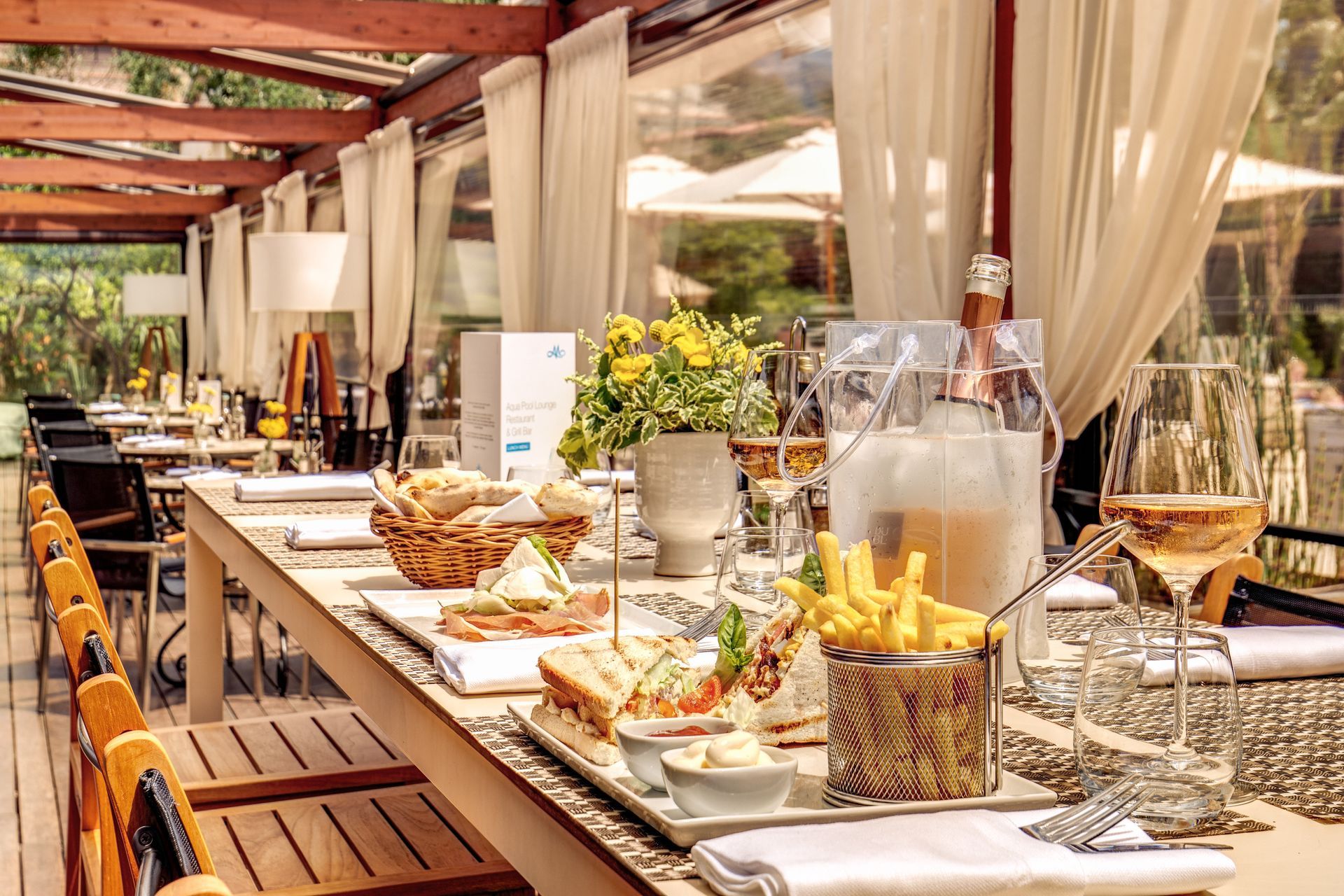 Elegant outdoor buffet table under a pergola, set with drinks, pastries, and flowers in a sunlit garden restaurant