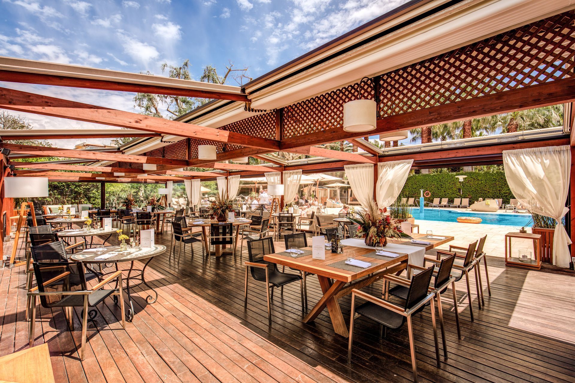 Open-air restaurant patio with wooden tables, lattice roof, and poolside view in warm sunlight