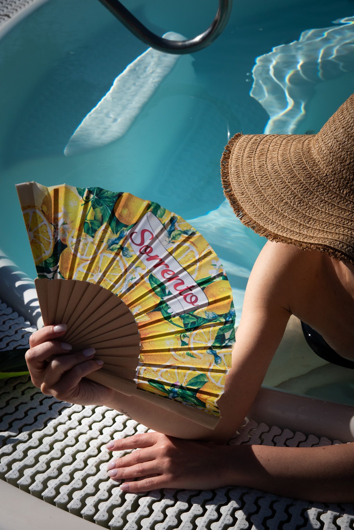 A person wearing a straw hat rests by a swimming pool while holding a yellow fan patterned with lemons.