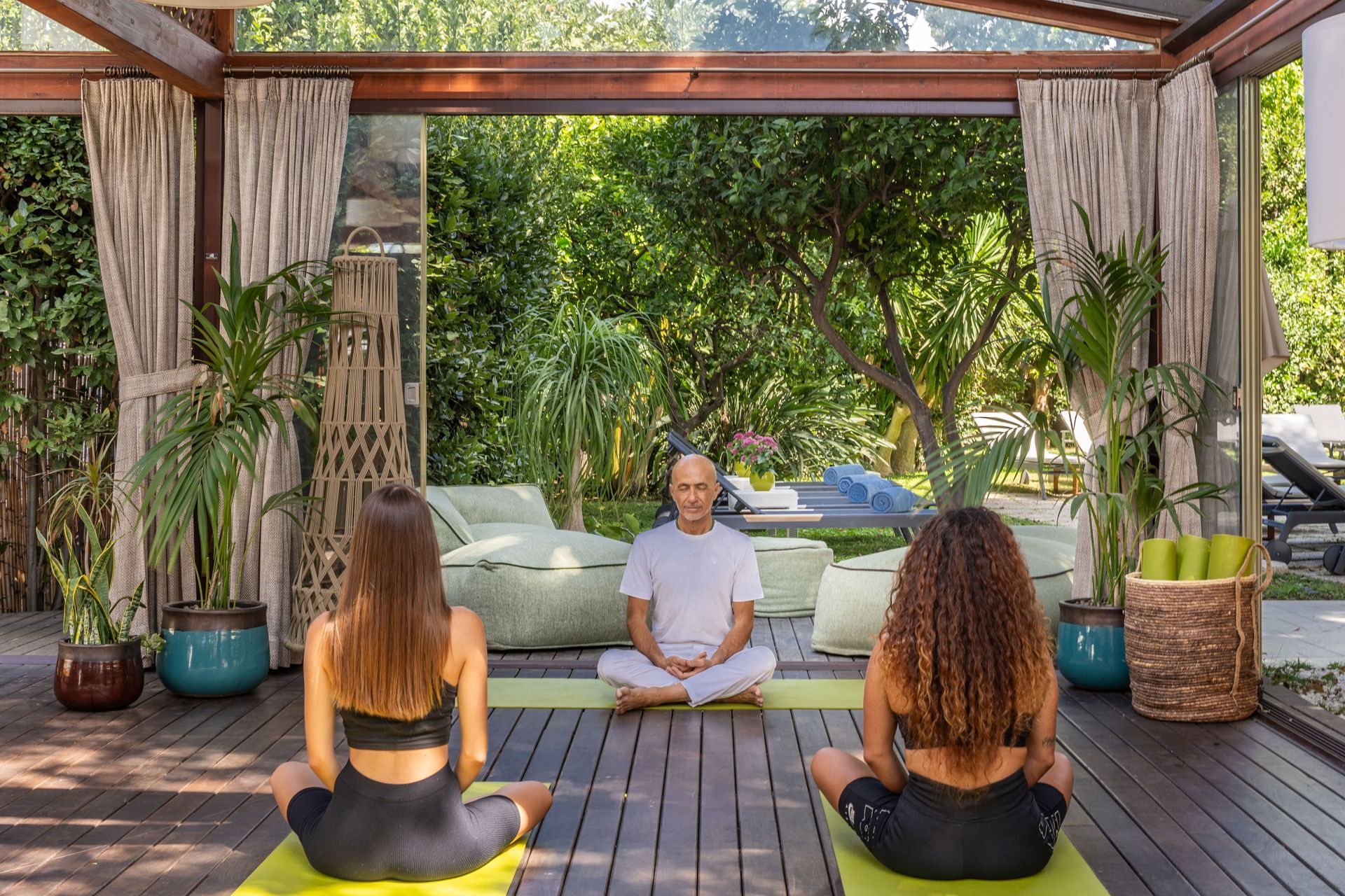 Two people and an instructor sit in meditation poses on mats on a wooden deck in front of a lush garden.